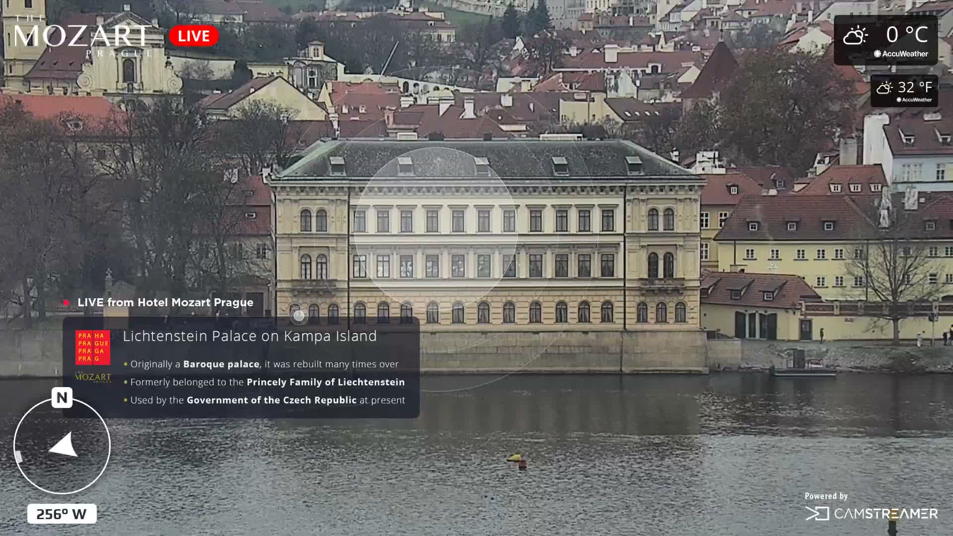 A grand, multi-story palace with many windows stands on the bank of a wide river, surrounded by other buildings and bare trees under an overcast sky.