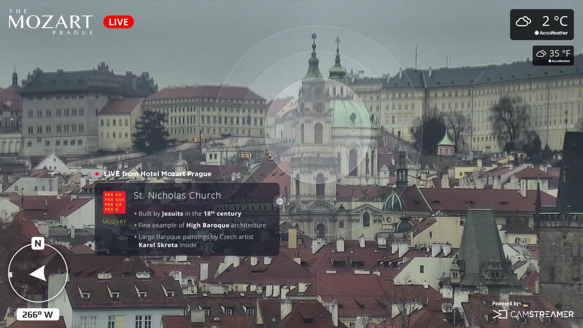 A grand, multi-story palace with many windows stands on the bank of a wide river, surrounded by other buildings and bare trees under an overcast sky.