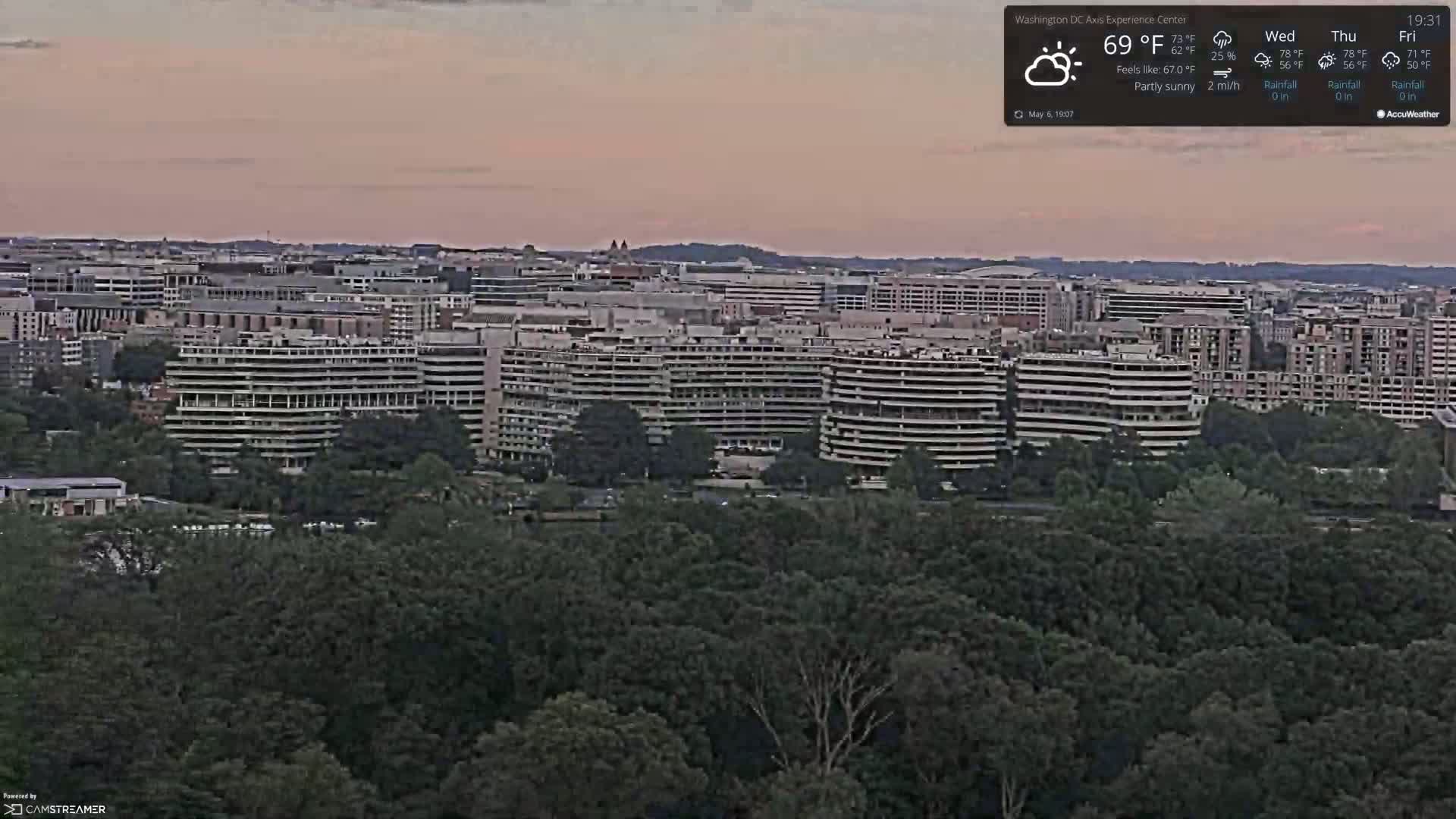 A partly sunny evening view of a cityscape with numerous high-rise buildings, nestled amongst a lush green forest.