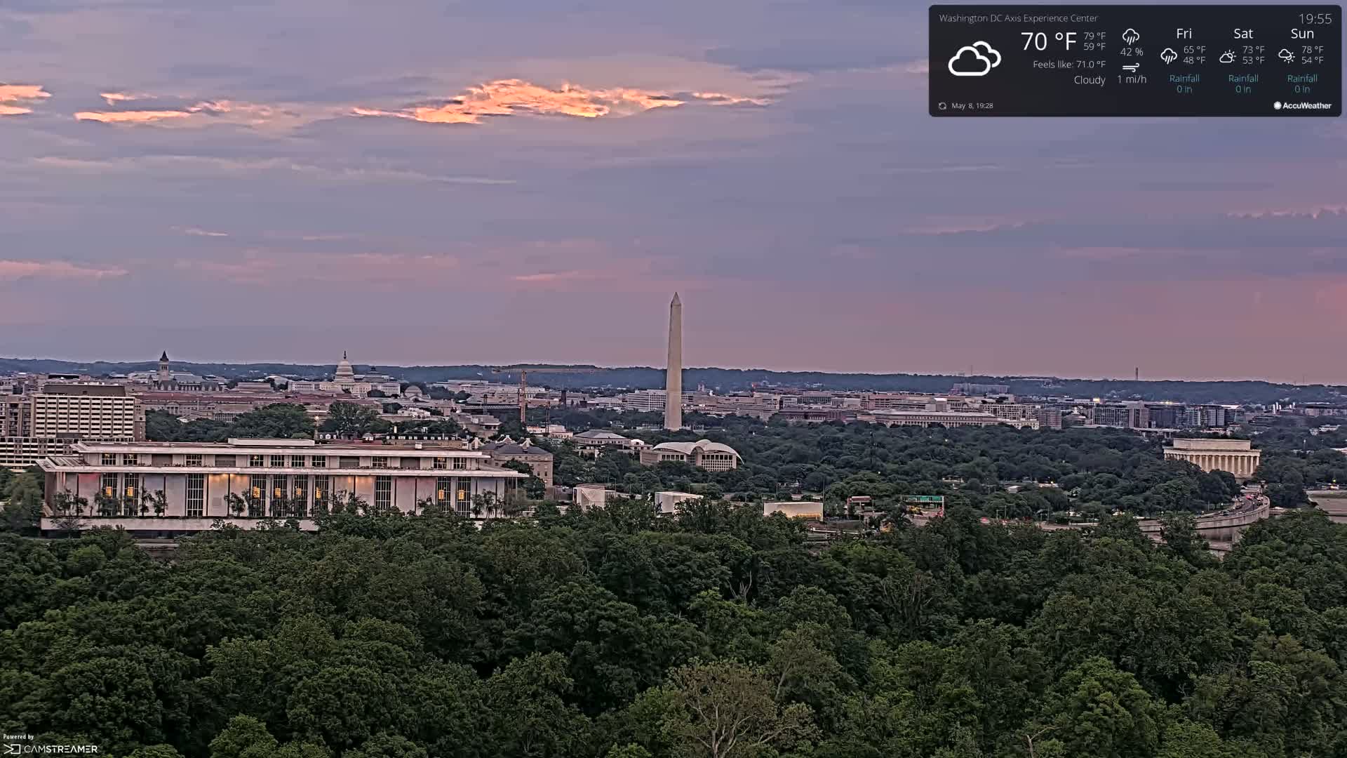 A cloudy, twilight view of Washington, D.C., shows the Washington Monument and other landmarks surrounded by green trees and buildings.