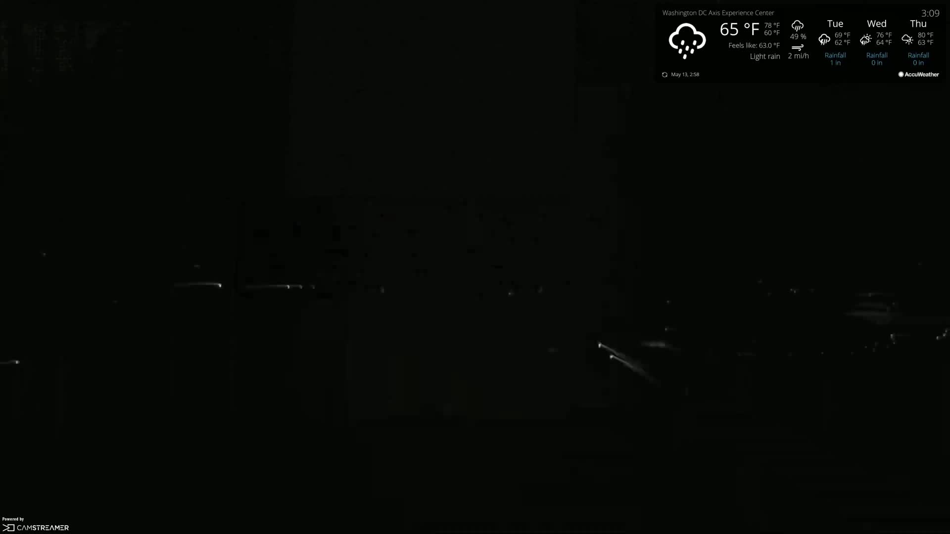 A nighttime, low-light view of a dark scene with light rain and faint, distant lights.