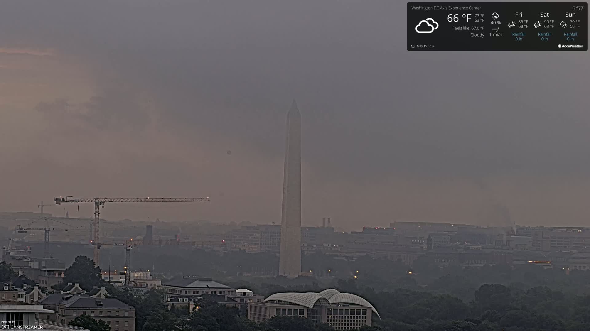 A hazy, cloudy cityscape with the Washington Monument prominently visible in the center, featuring construction cranes in the foreground.