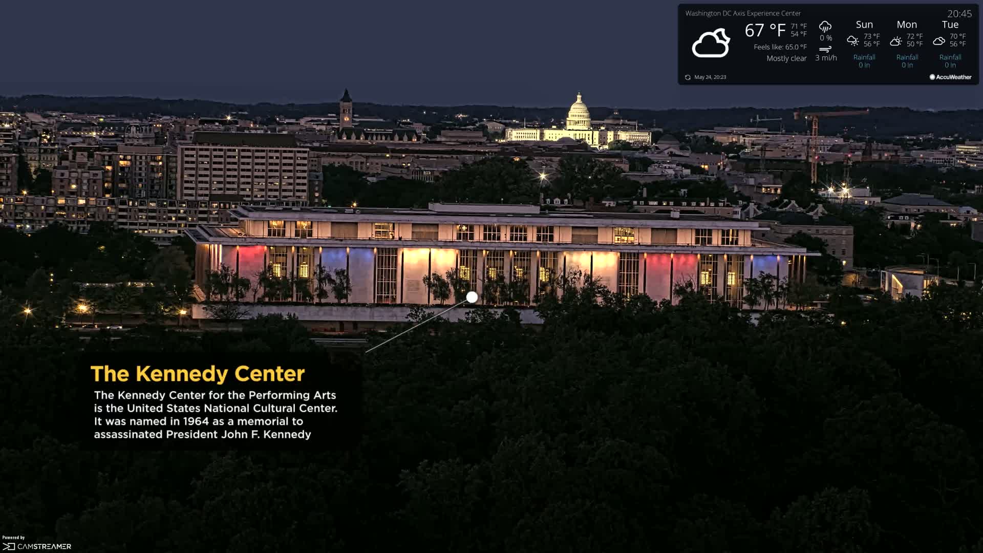The illuminated Kennedy Center is shown at night, under mostly clear skies, with the Capitol building visible in the distance.