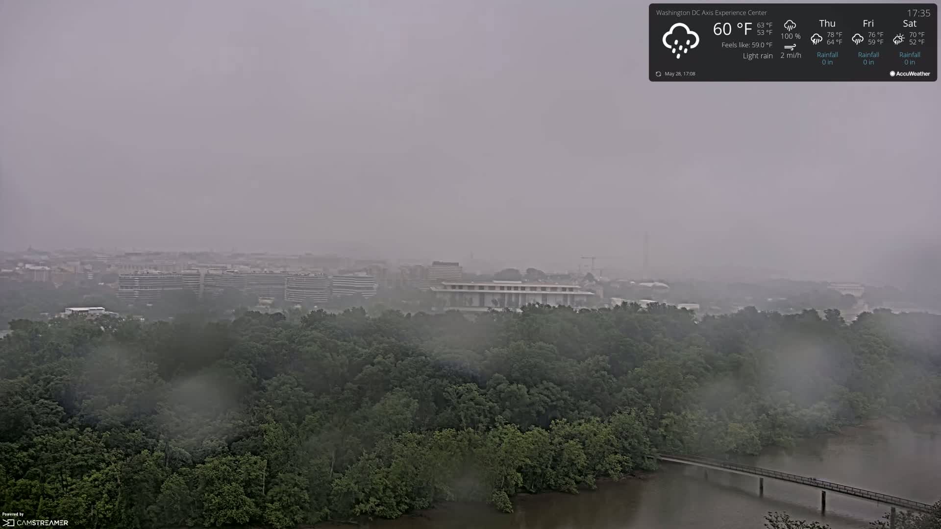 A hazy, light rain falls over a city skyline and lush green trees bordering a river with a small footbridge.