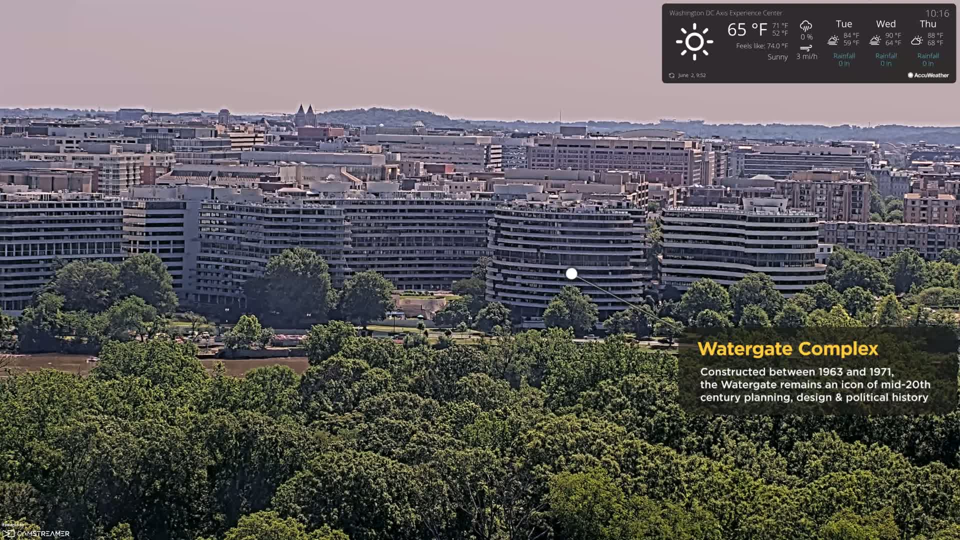 A sunny, high-angle view of the Watergate Complex in Washington, D.C., situated among lush green trees and overlooking a body of water, with a cityscape in the background.