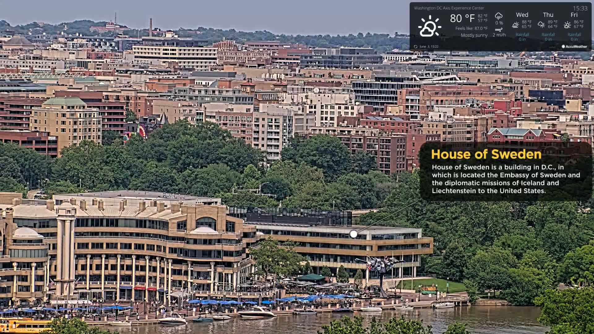 A large, tan building situated on a waterfront, surrounded by lush greenery and numerous other buildings in a city, under mostly sunny skies with a light breeze.