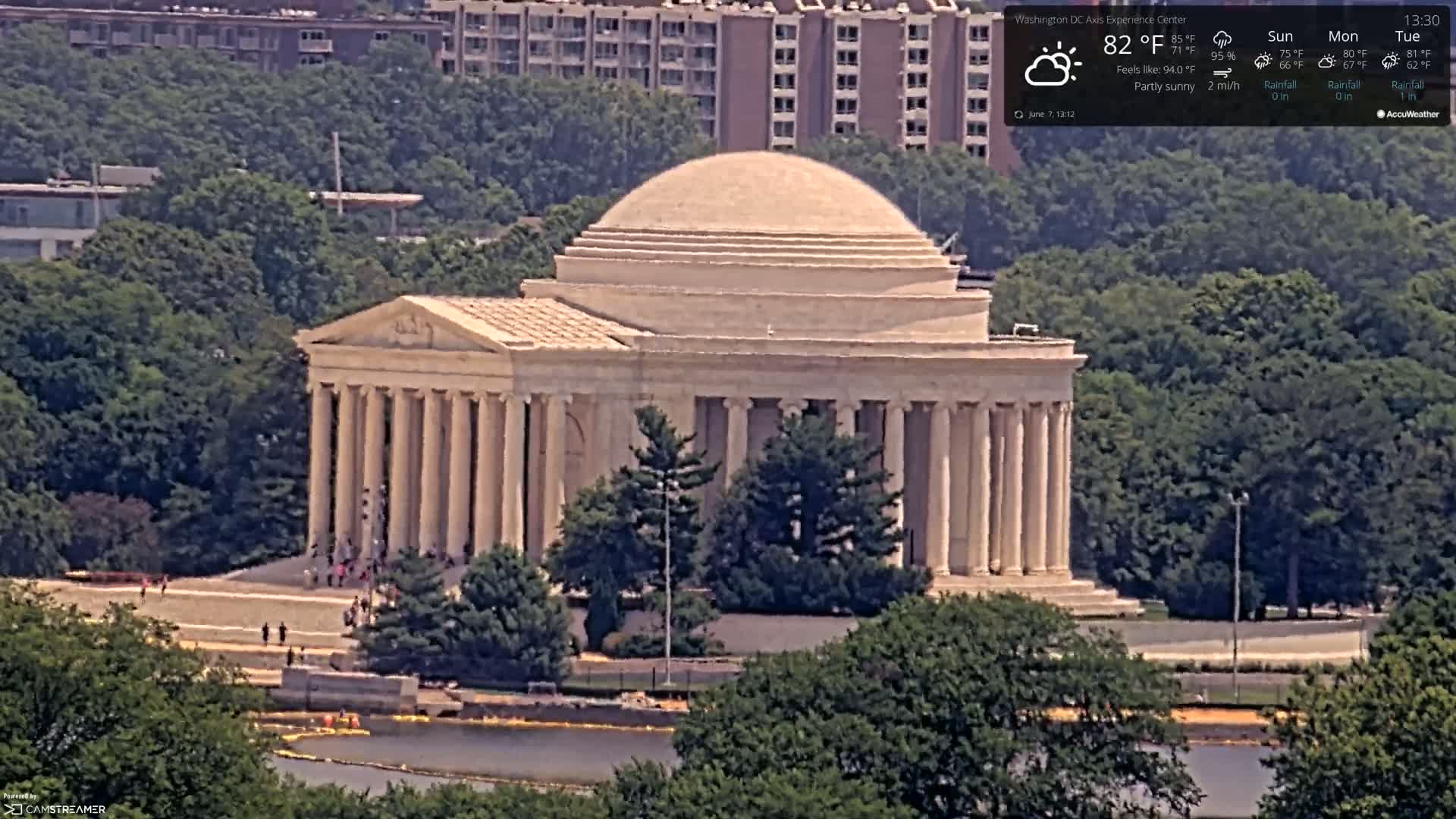 A large, classical-style building with a dome sits on a landscaped area surrounded by green trees on a partly sunny day.