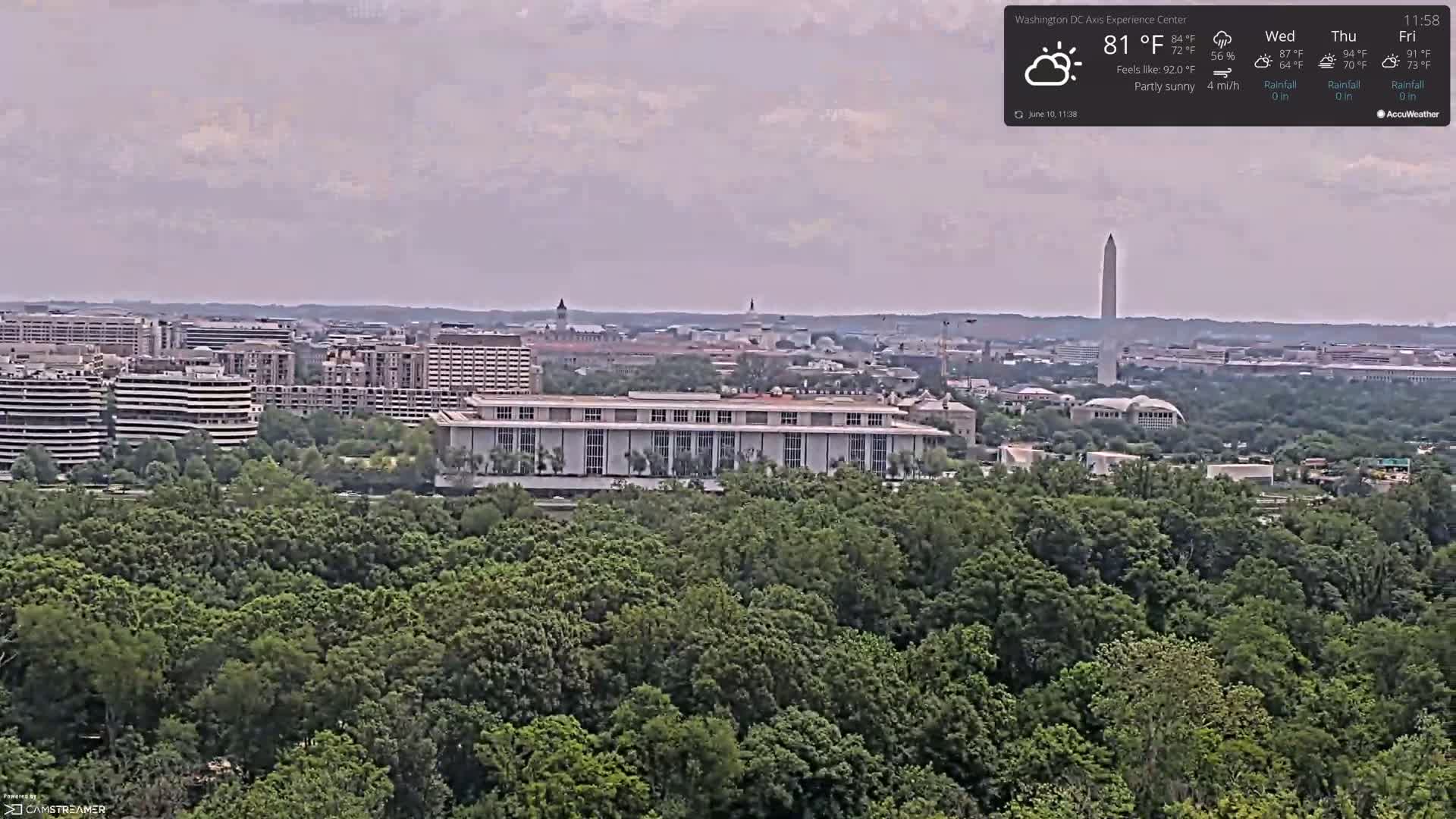 A partly sunny view from above a green forest shows a cityscape including the Washington Monument and numerous buildings under a hazy sky.