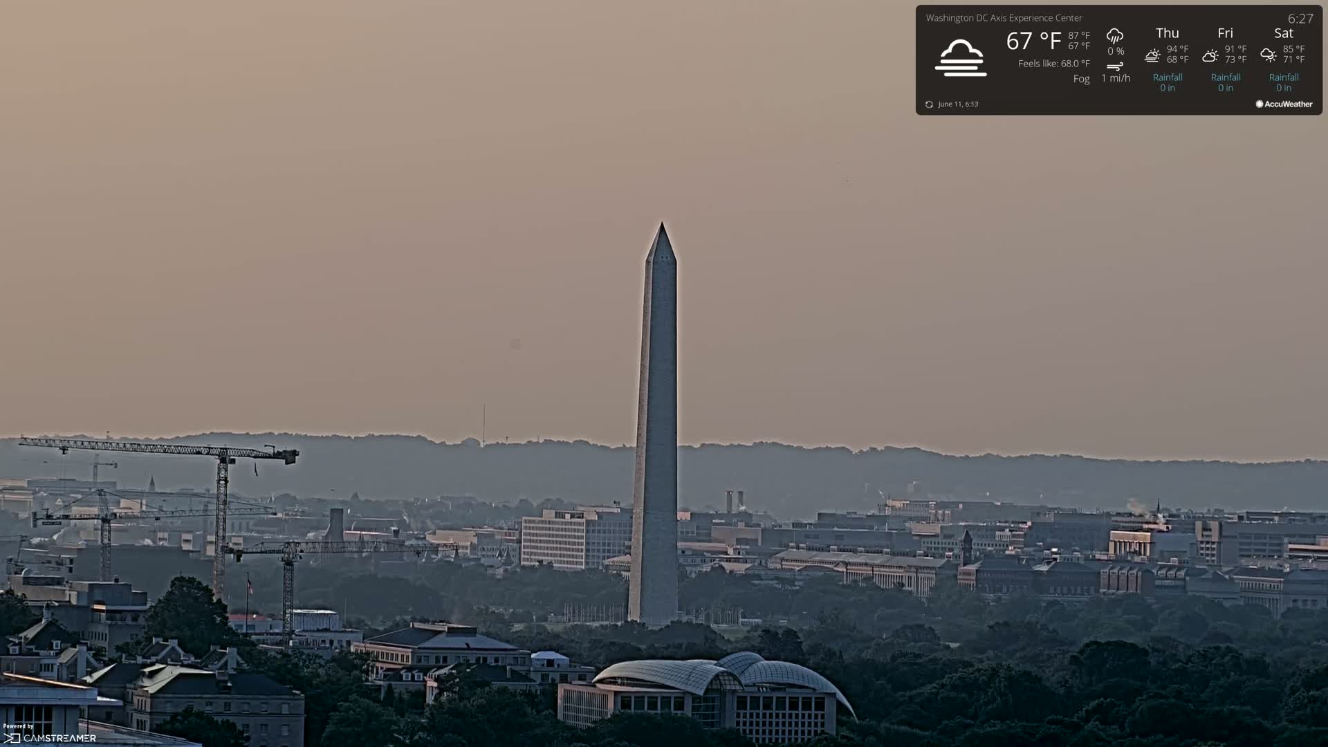 A hazy, foggy cityscape features the Washington Monument, construction cranes, and numerous buildings under a pale, dusky sky.