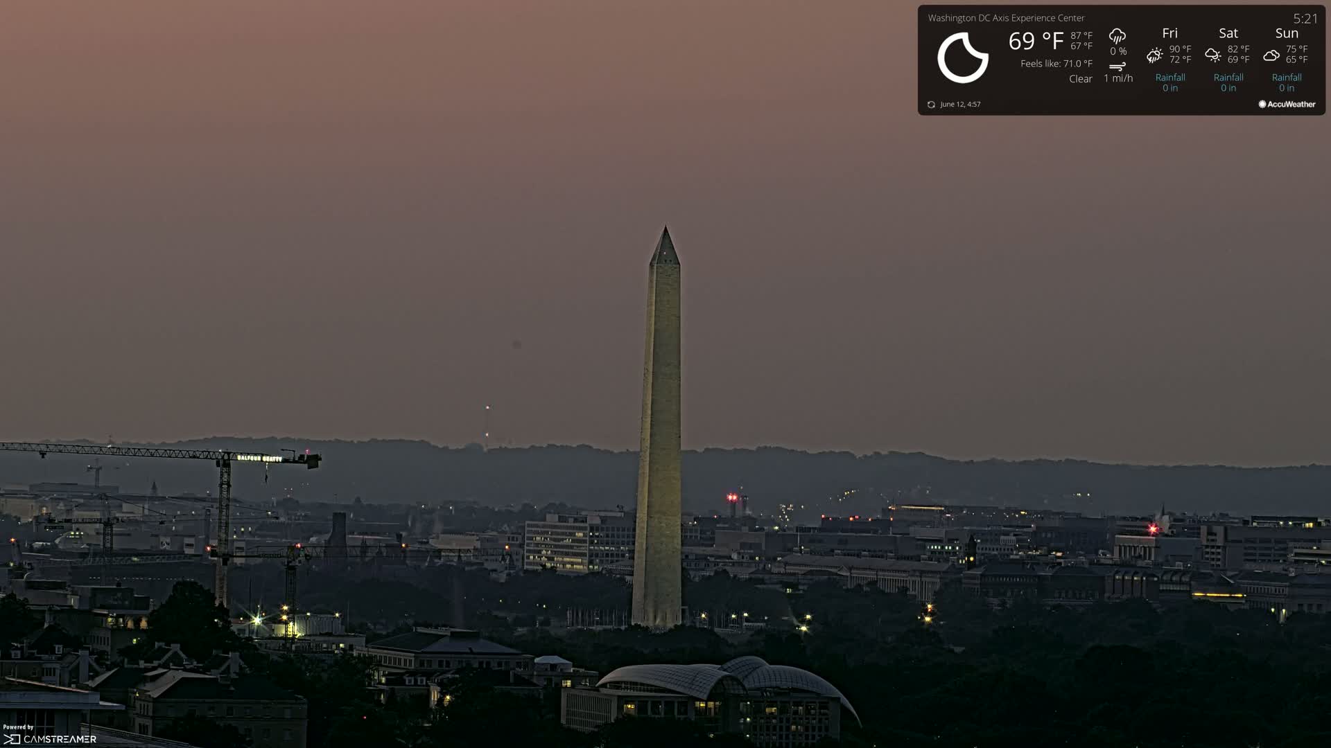 A twilight view of the Washington Monument, illuminated against a dusky sky, with a city skyline and construction cranes visible in the foreground.