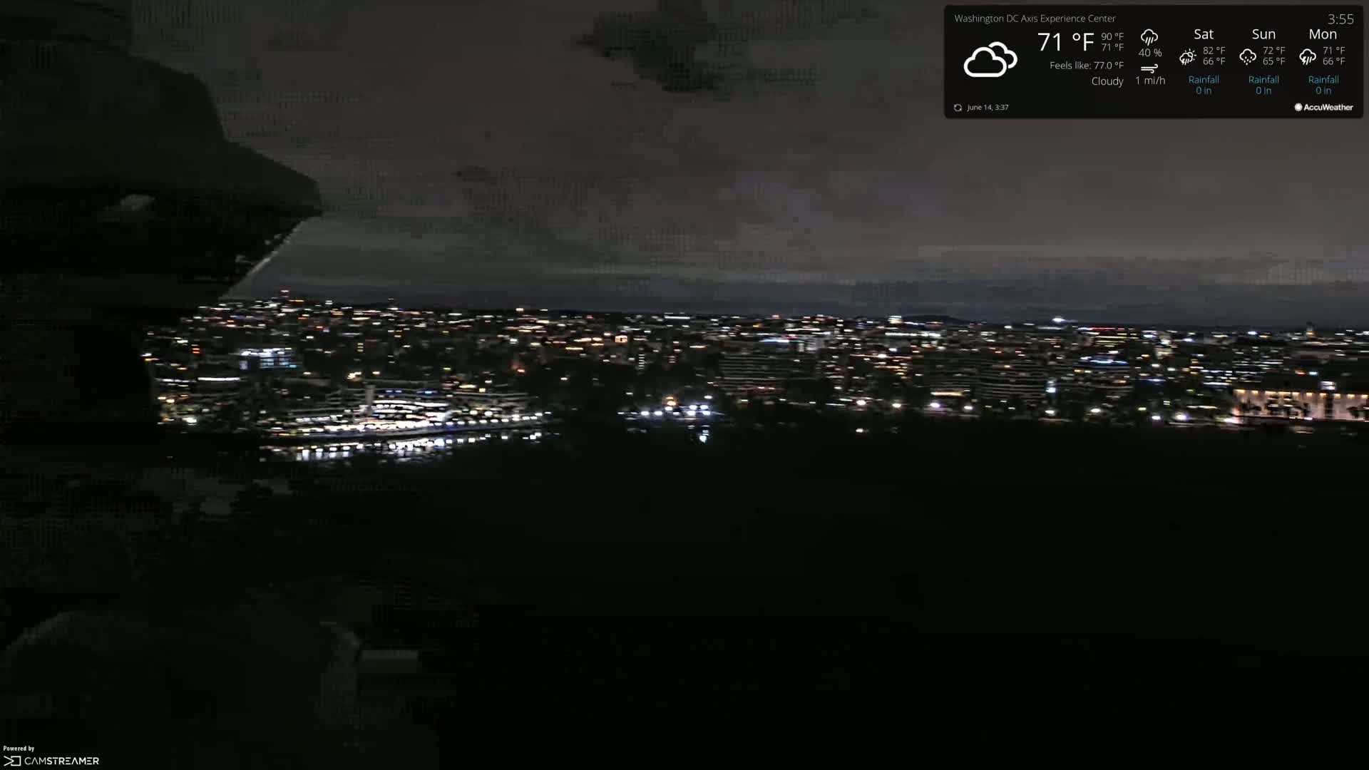 A nighttime, cloudy view of a city illuminated by numerous lights, with a body of water visible in the foreground.