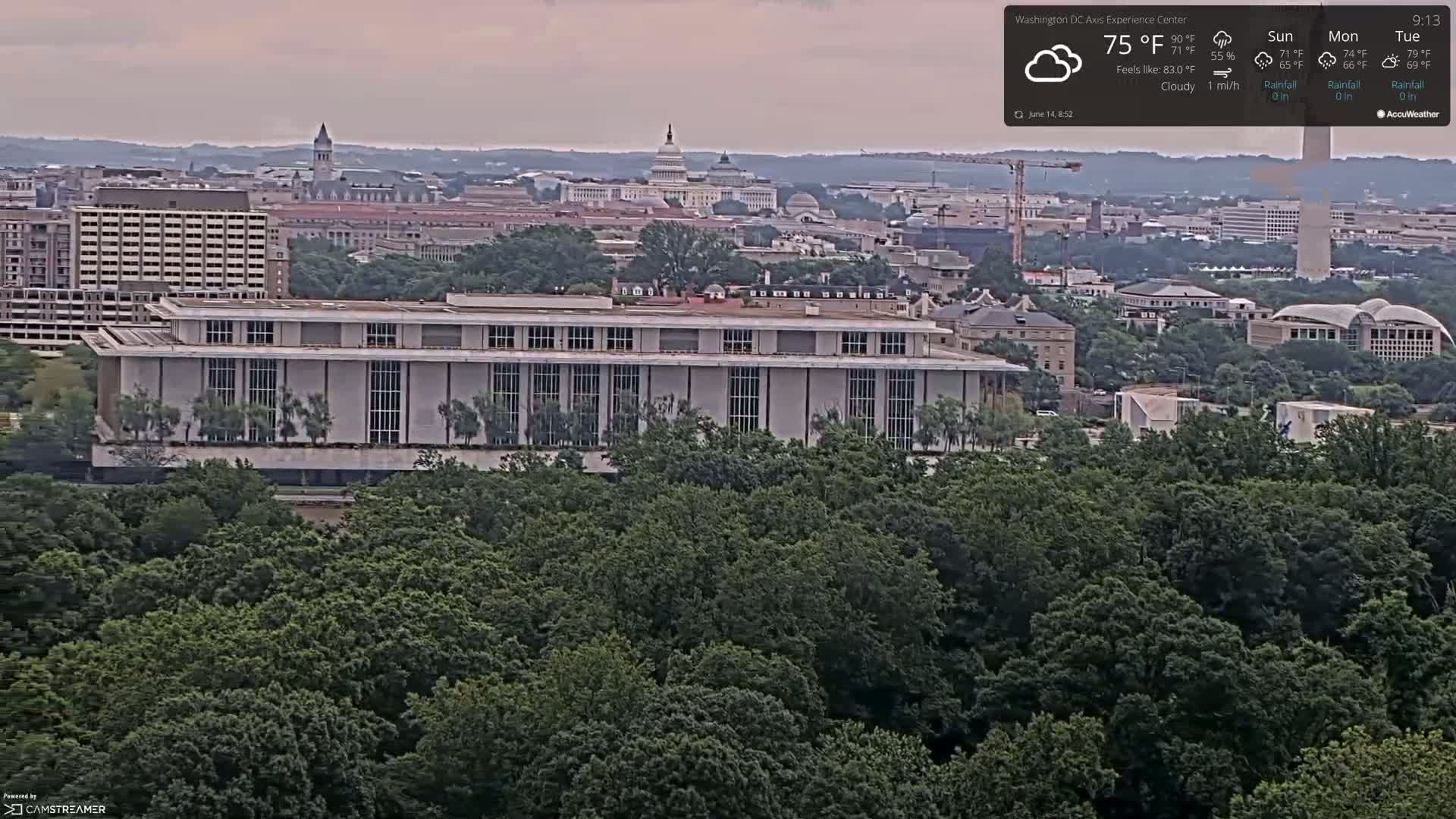 A long, low building sits in a lush green forest overlooking a city skyline with the Capitol building visible in the distance under cloudy skies.