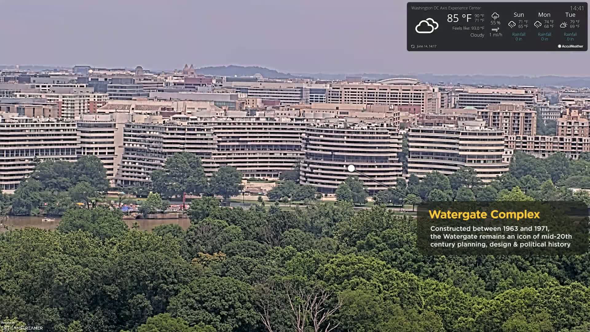 A long, low building sits in a lush green forest overlooking a city skyline with the Capitol building visible in the distance under cloudy skies.