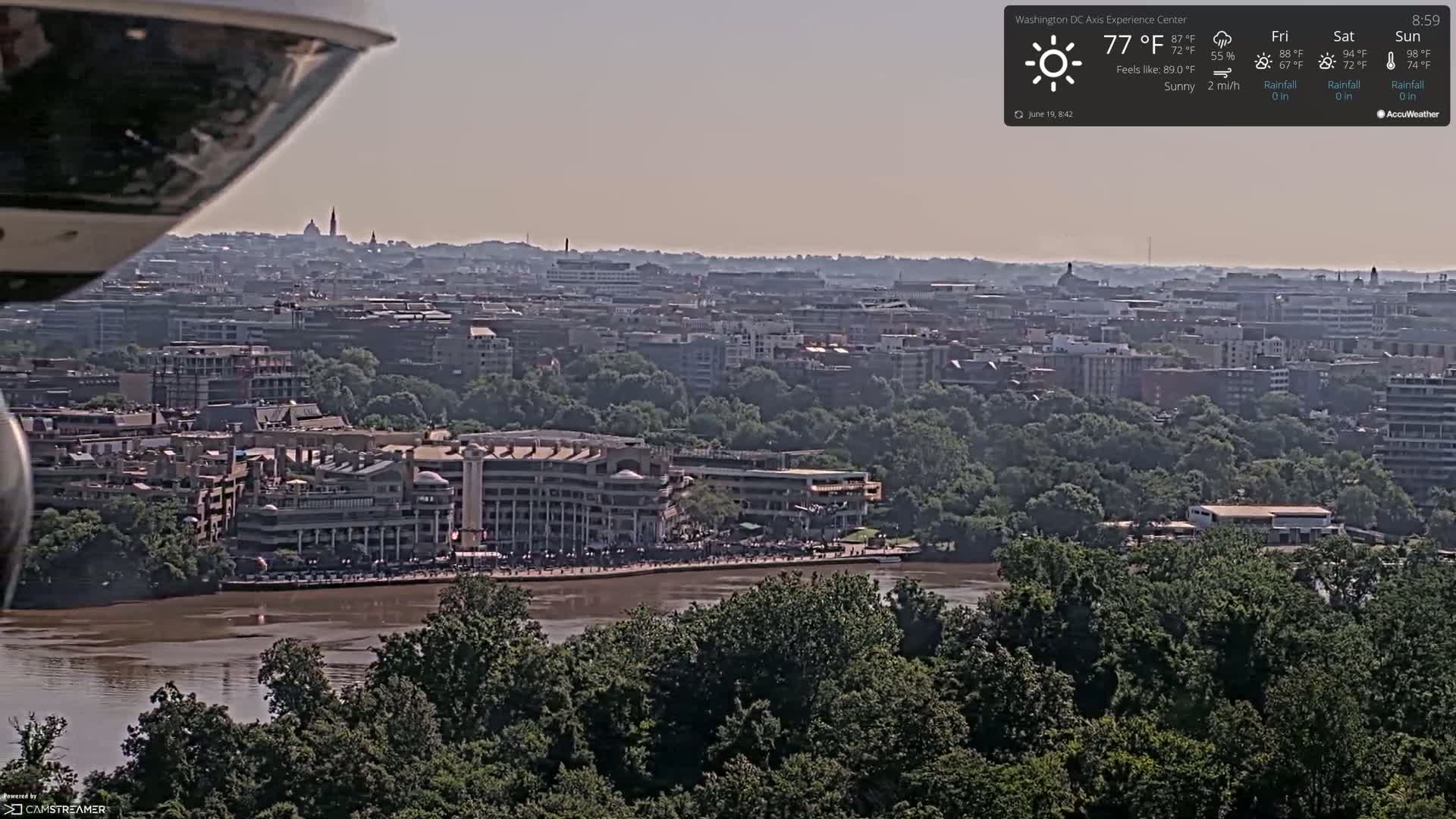 A sunny, hazy view of a city sprawling along a brown river, with a large building complex situated near the water's edge.