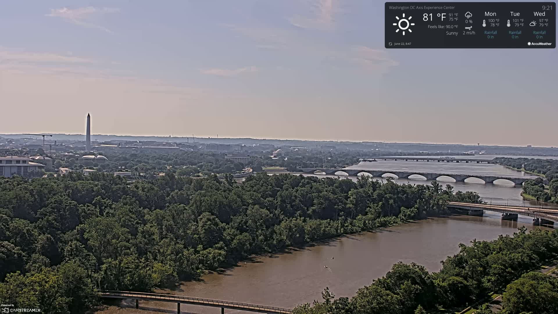 A sunny, high-angle view shows a wide river with two bridges, lush green trees lining its banks, and the Washington Monument visible in the distance.