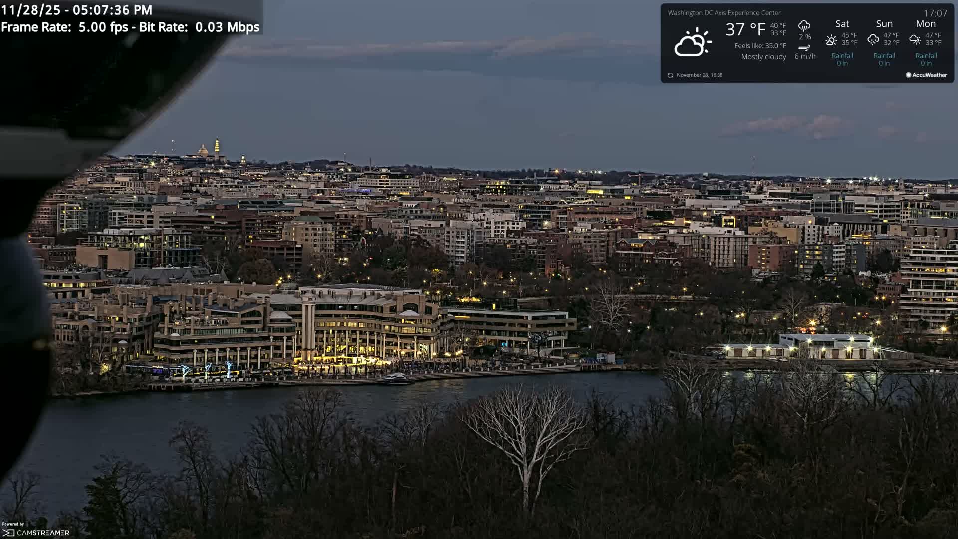 A wide panoramic view of a city at dusk, with a river in the foreground bordered by brightly lit buildings, and a dense cityscape stretching into the background under a cloudy sky.