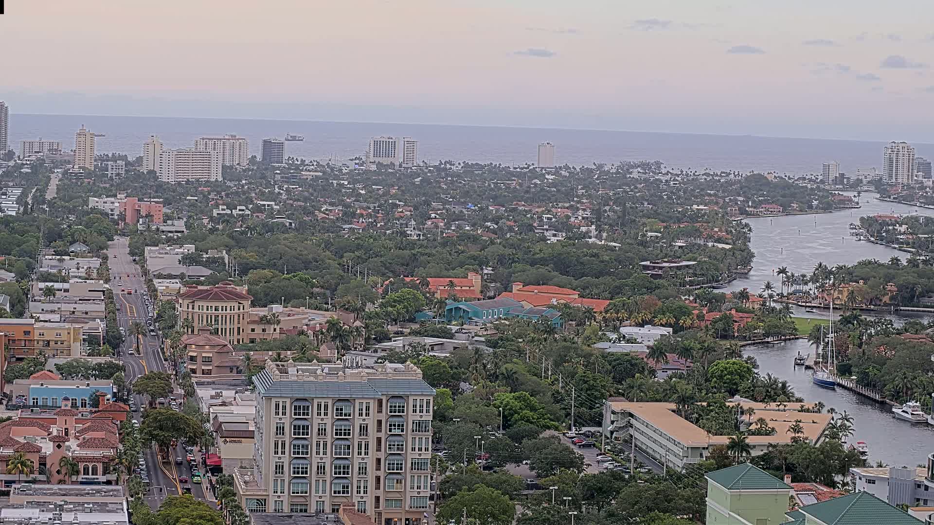 An aerial view shows a coastal city with a waterway winding through a densely populated area of low-rise buildings and lush vegetation, under an overcast sky.