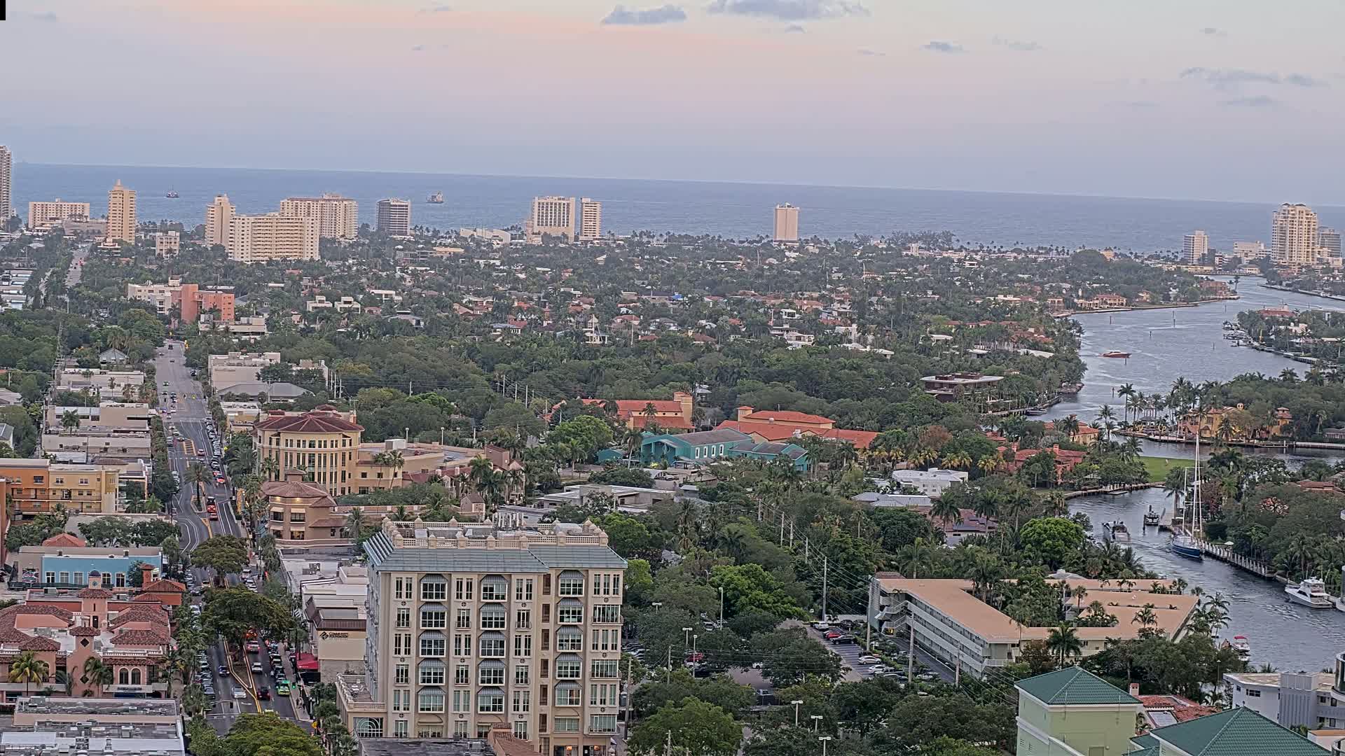 A coastal city with a waterway winds through a dense, lush residential area under a partly cloudy sky, with the ocean visible in the distance.