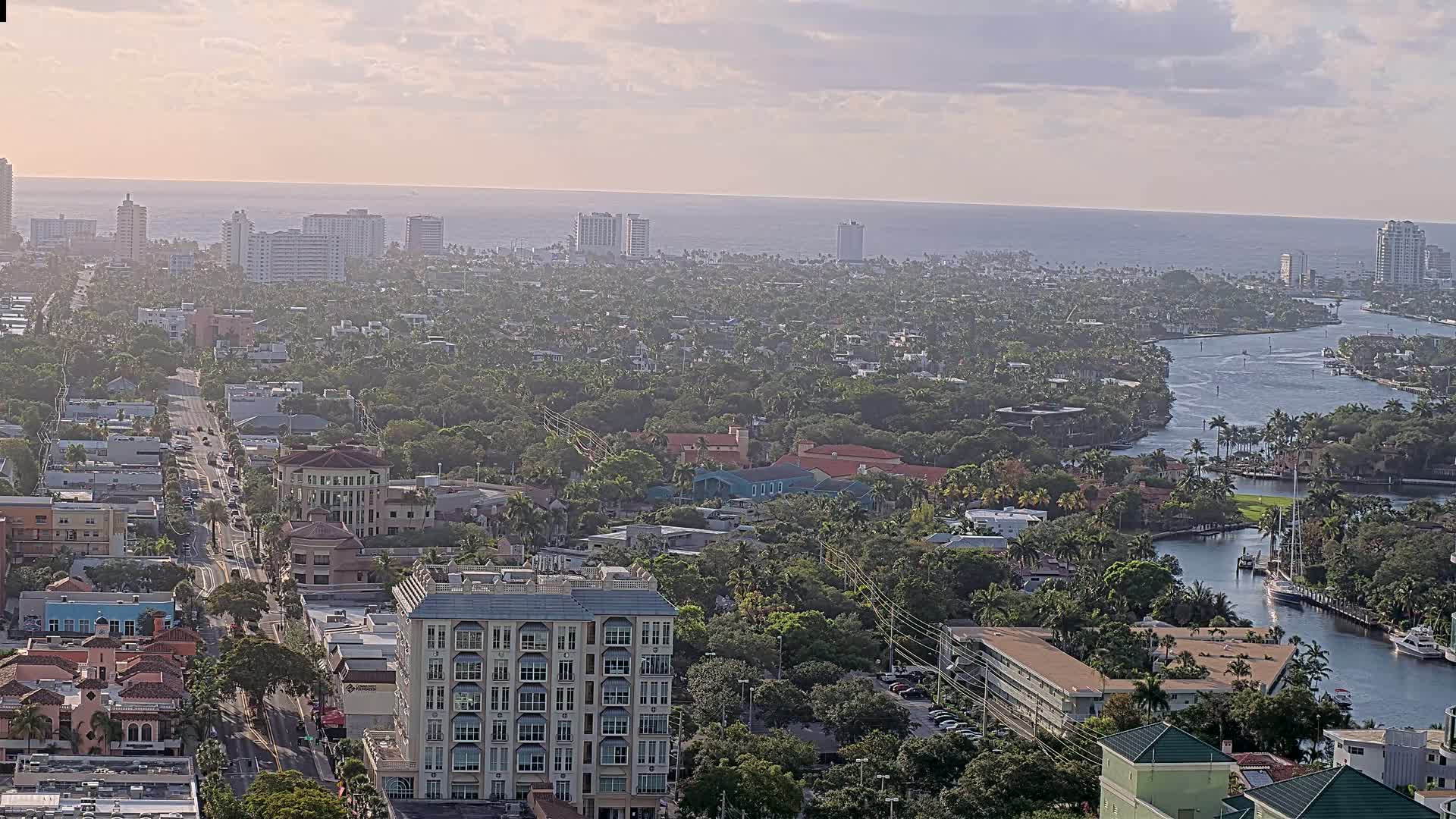 An aerial view shows a coastal city with buildings, palm trees, waterways, and a hazy, sunny sky.