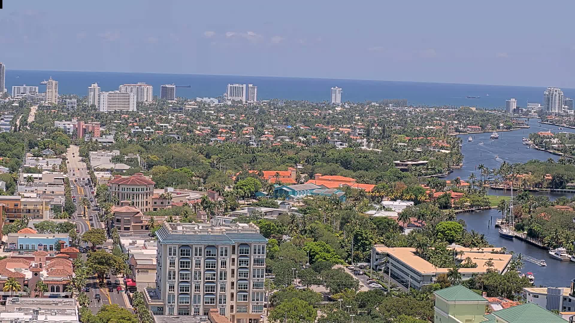 A coastal city with a waterway winds through a neighborhood of lush green trees and terracotta-roofed buildings, under a bright, sunny sky and calm ocean in the distance.