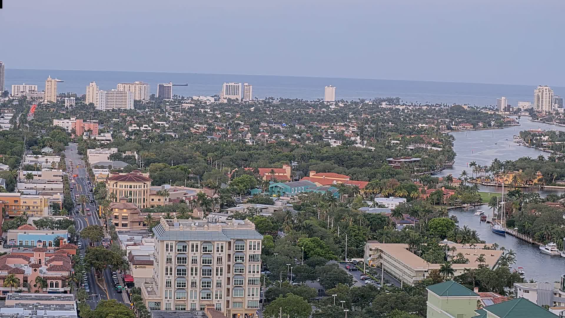 A coastal city with a calm ocean in the background, featuring a main road lined with buildings and a waterway winding through a lush, green residential area.
