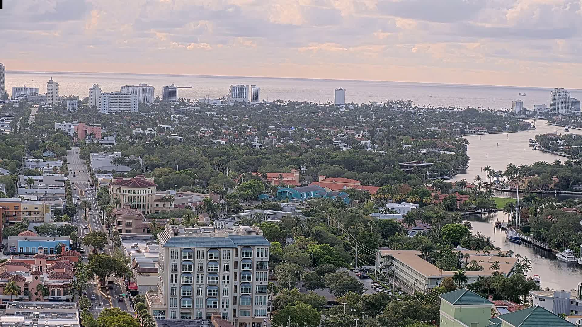 An aerial view of a coastal city with a calm ocean in the background under a cloudy sky shows a long road lined with buildings and a waterway winding through lush green landscaping.