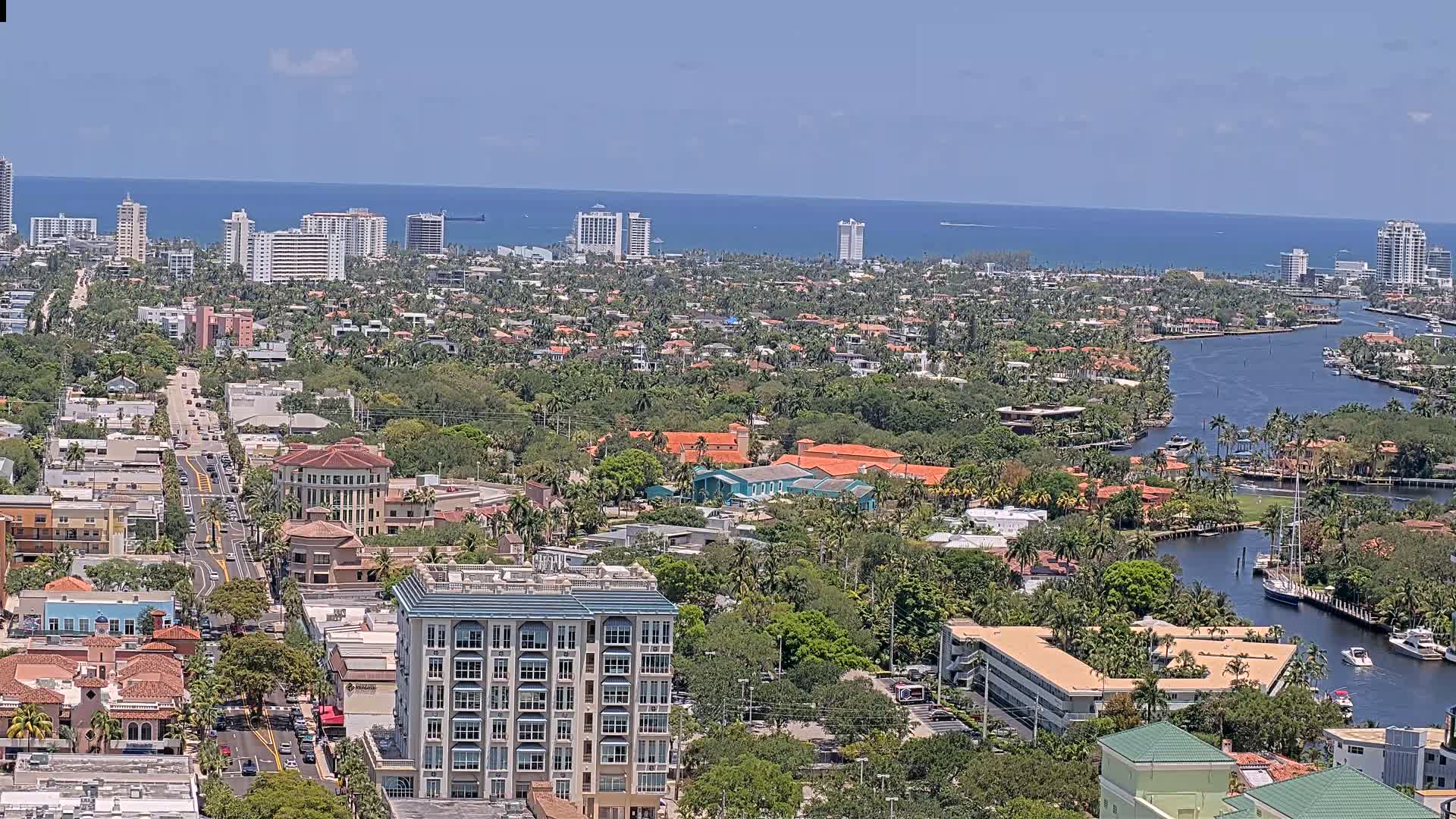 A coastal city with a waterway winds through a residential area of low-rise buildings and lush vegetation under a clear blue sky.