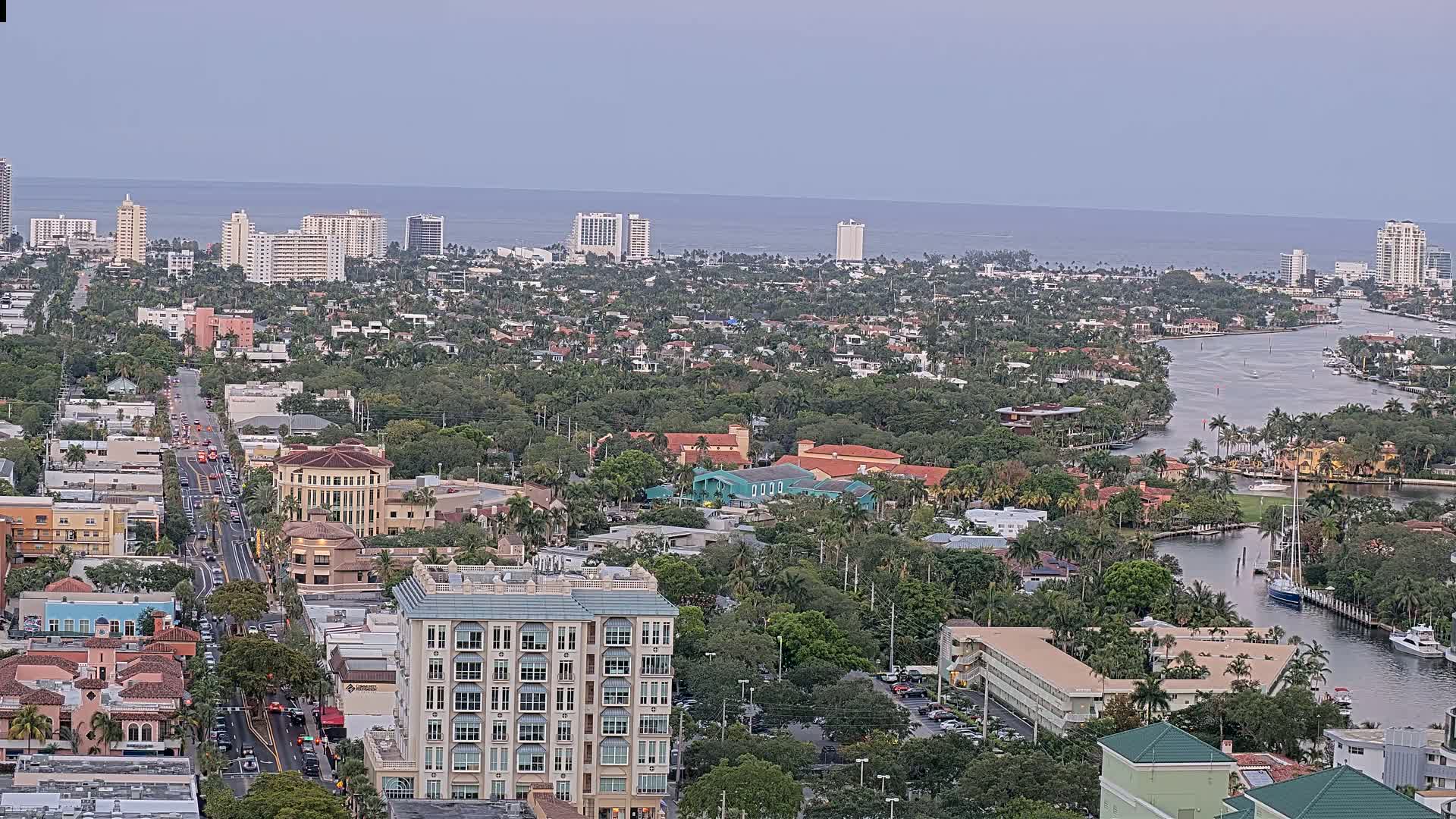 An aerial view shows a coastal city with numerous buildings, lush green vegetation, and a waterway winding through the residential and commercial areas under a cloudy sky.