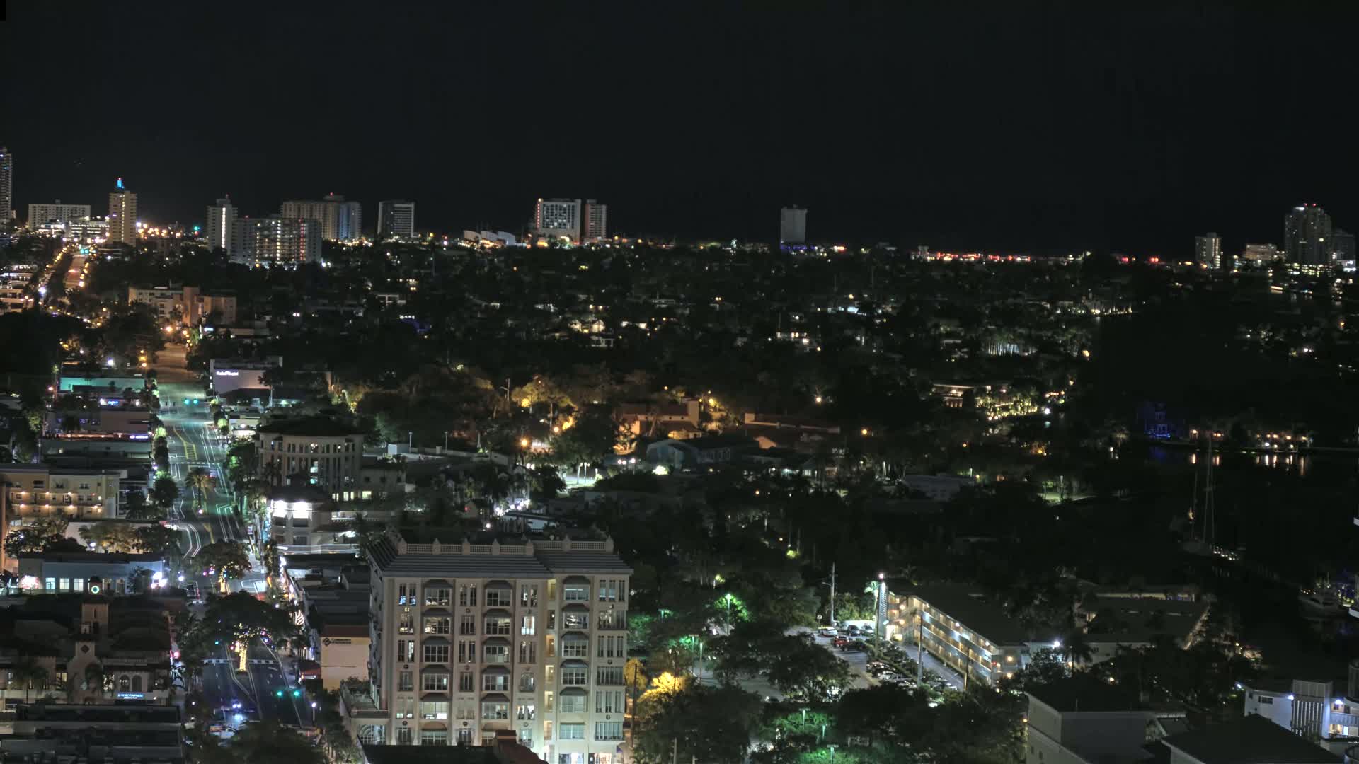 A nighttime aerial view shows a city with illuminated streets and buildings, surrounded by dark, tree-covered areas.