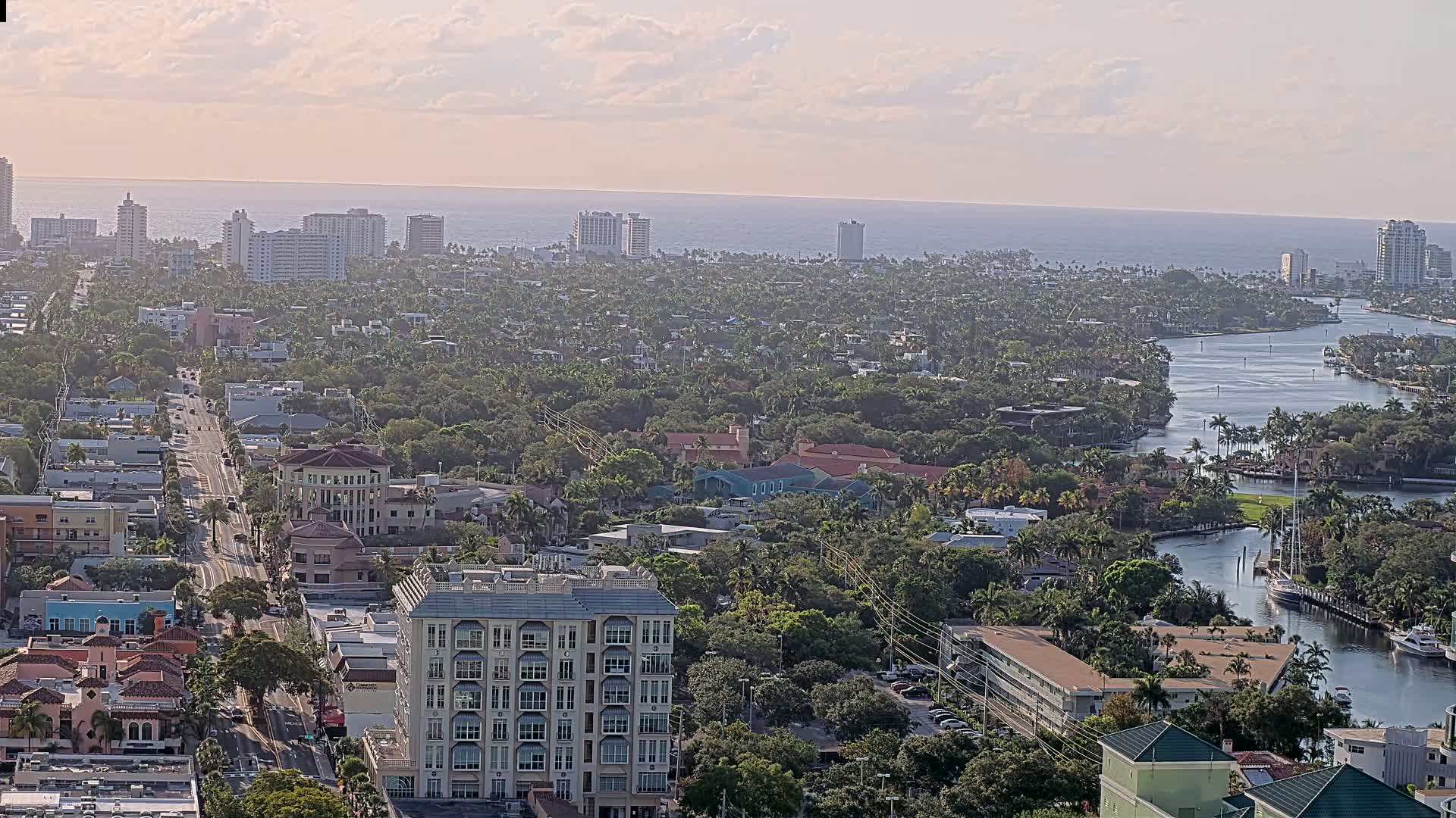 An aerial view shows a coastal city with a waterway winding through a dense, lush landscape under a mostly clear sky.