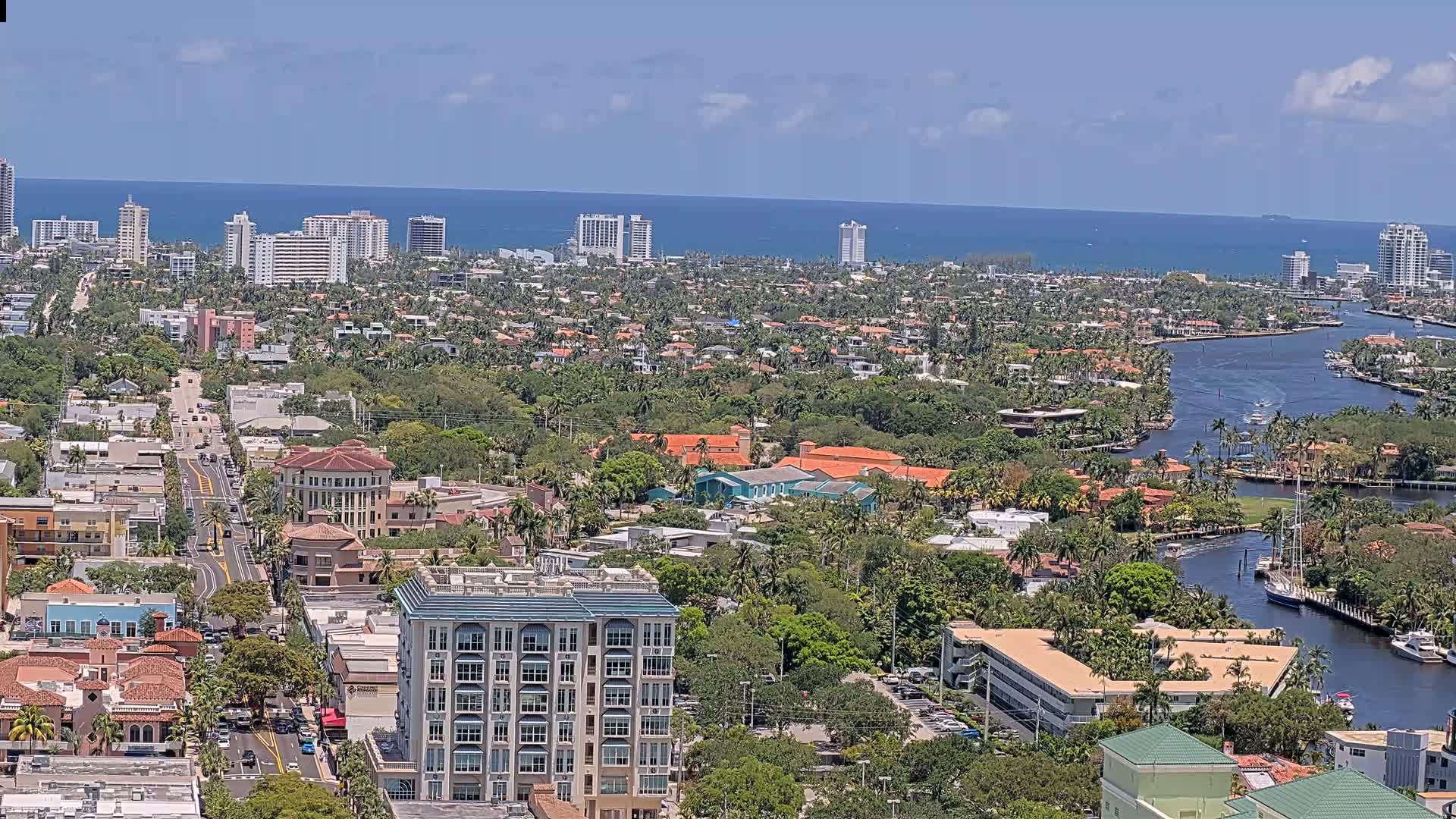 An aerial view on a sunny day shows a coastal city with numerous buildings, lush greenery, a waterway, and a distant ocean.
