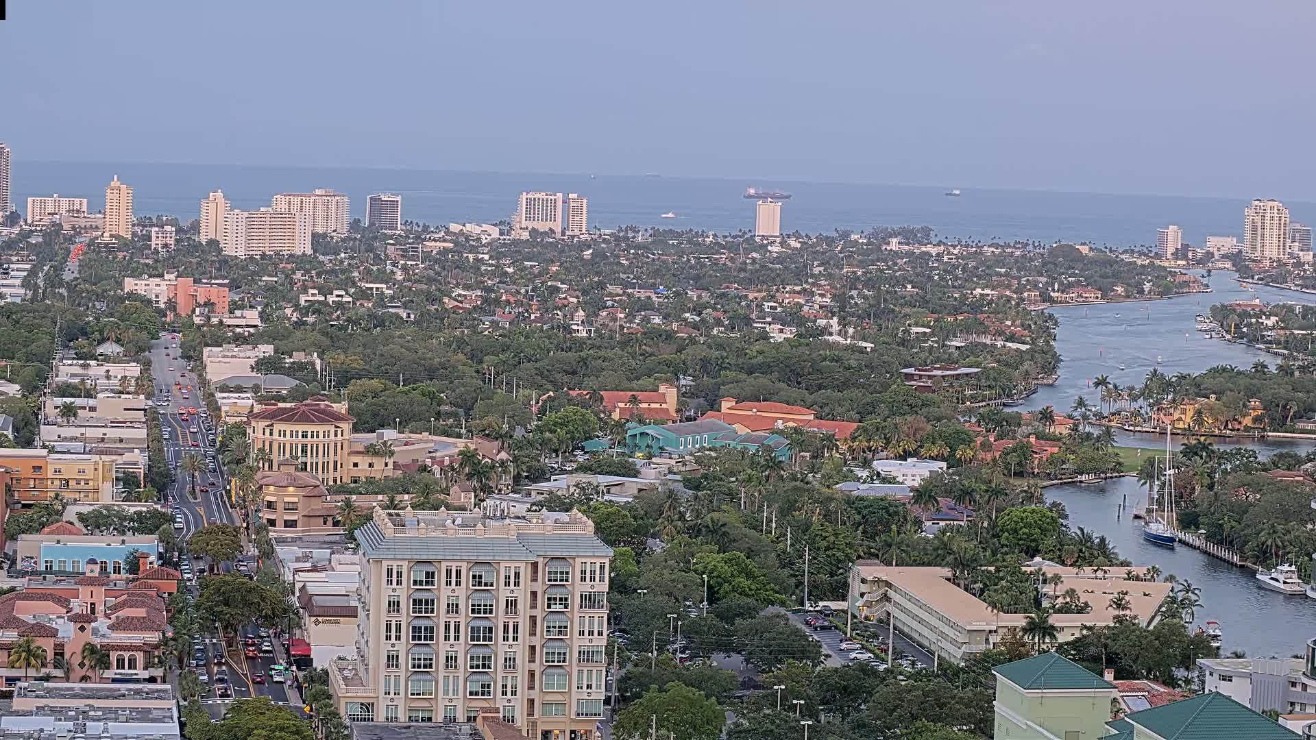 An aerial view of a coastal city on a partly cloudy day, showing a network of waterways winding through densely populated residential areas and high-rise buildings, with a calm ocean visible in the distance.