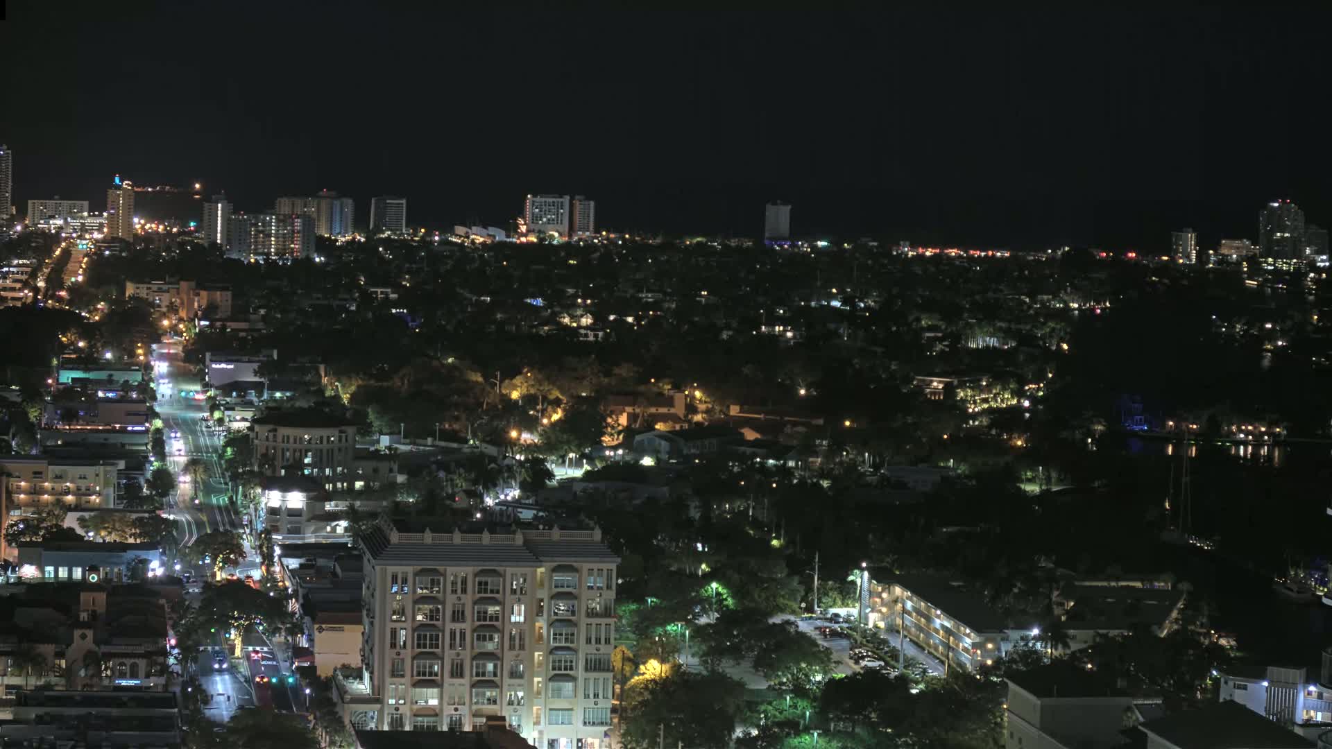 A nighttime aerial view shows a city with illuminated streets and buildings, surrounded by dark, tree-covered areas.