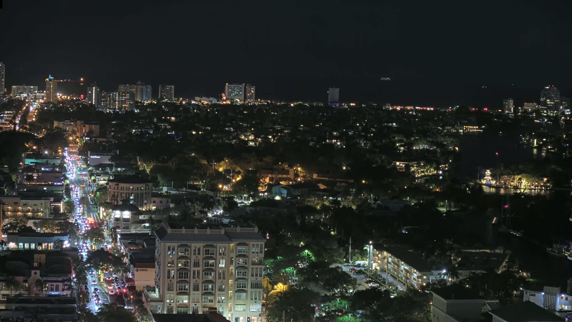 A nighttime aerial view shows a city with a main road congested with traffic, surrounded by residential areas and a waterfront with boats.