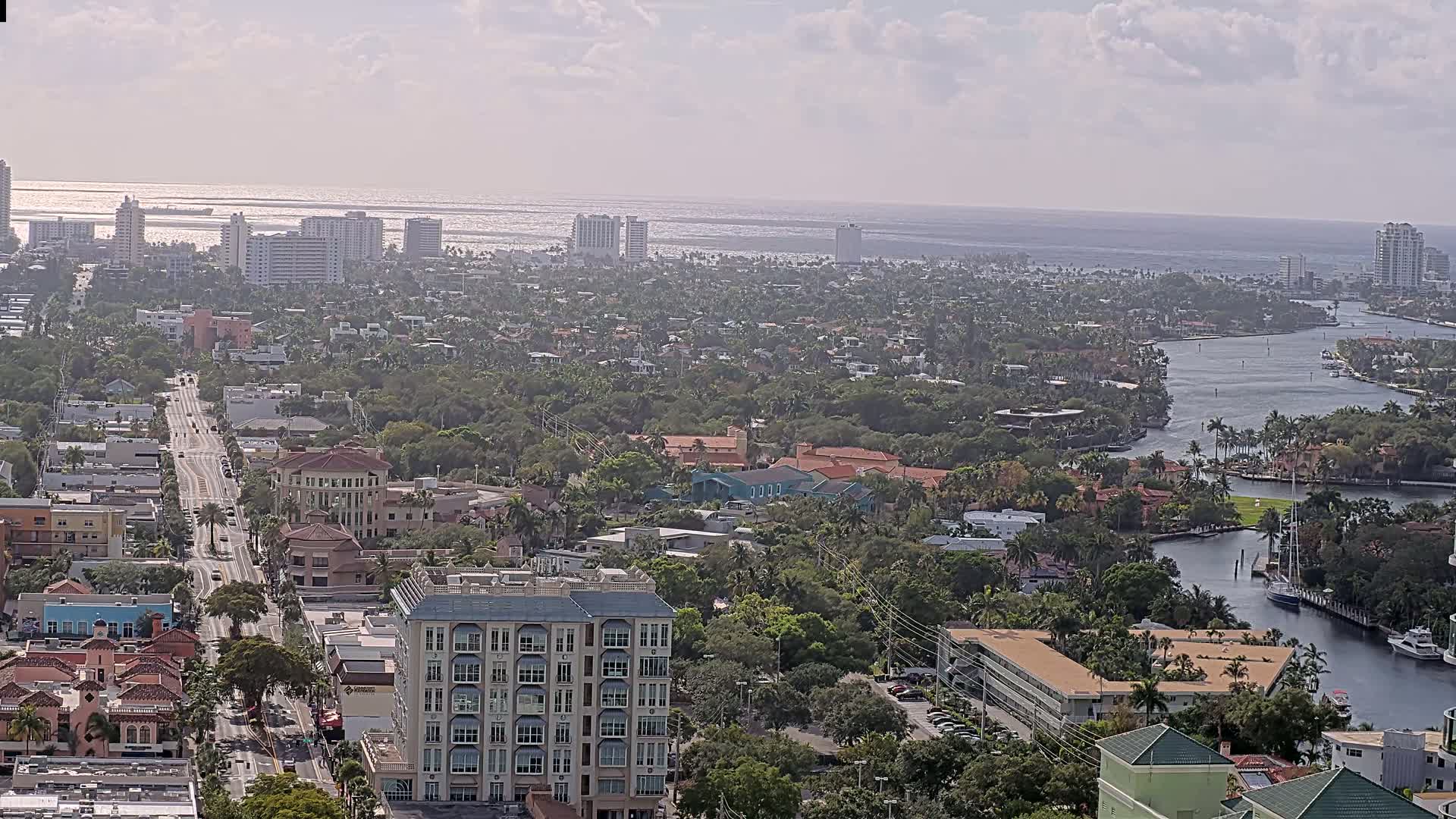 A coastal city with a waterway winding through a dense, green residential area, under partly cloudy skies.