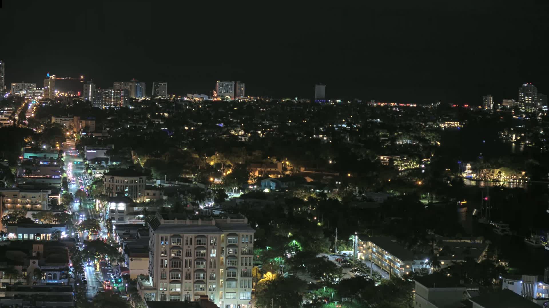 A nighttime aerial view shows a city with numerous lit buildings and streets, some taller buildings in the distance, and dark green areas interspersed throughout.