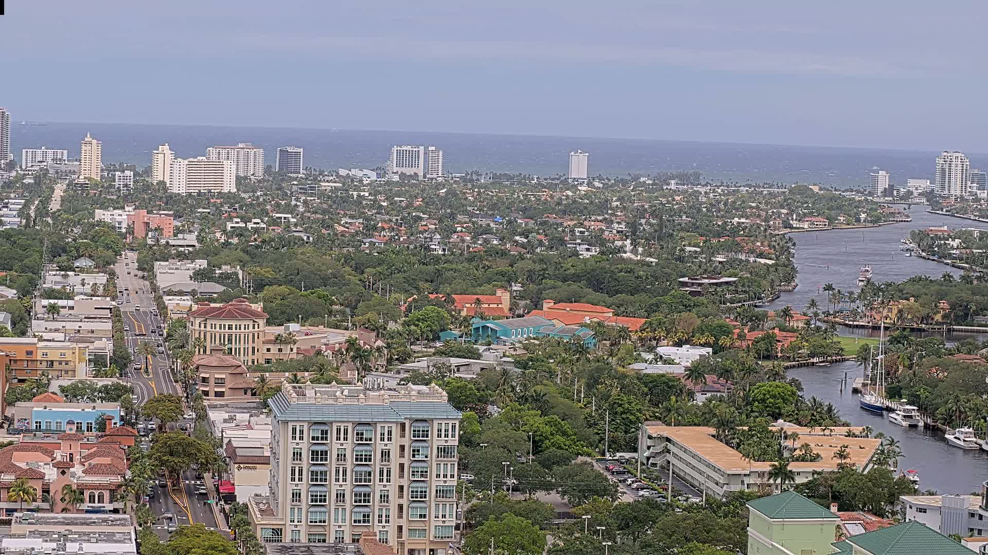 An aerial view of a coastal city on an overcast day, showing a network of waterways winding through a densely built-up area with numerous residential buildings and high-rises, bordered by lush greenery and the ocean in the distance.