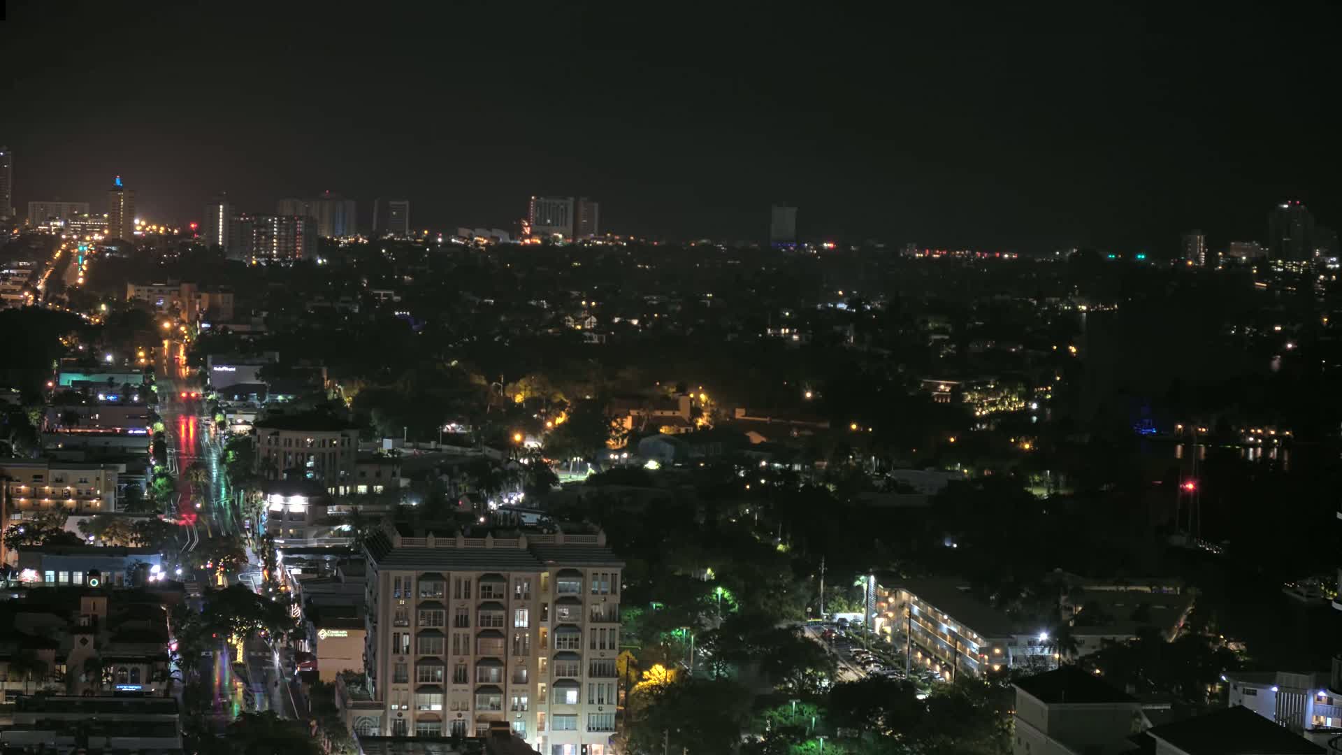 A rain-slicked city street cuts through a densely populated area at night, surrounded by low-rise buildings and taller structures in the distance, all illuminated under a dark sky.