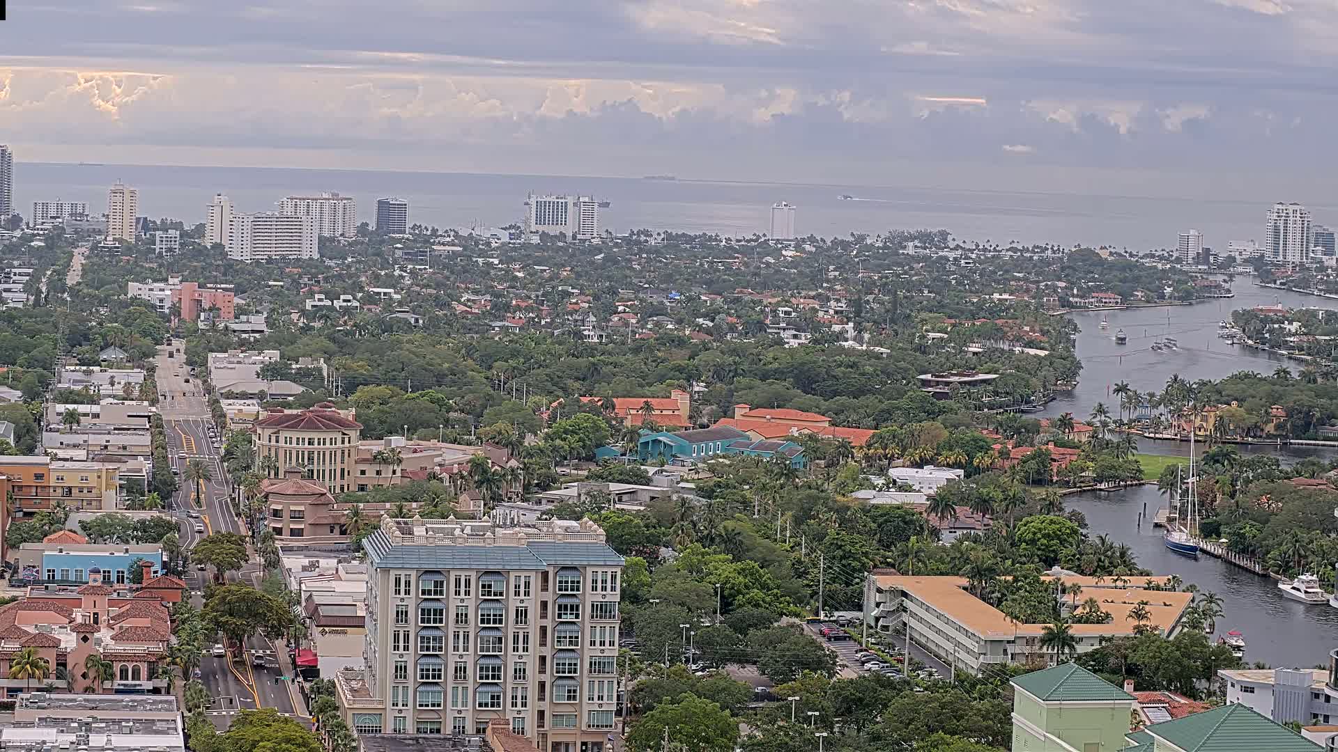 A coastal city with a waterway winds through a dense, tree-lined residential area under a cloudy sky.