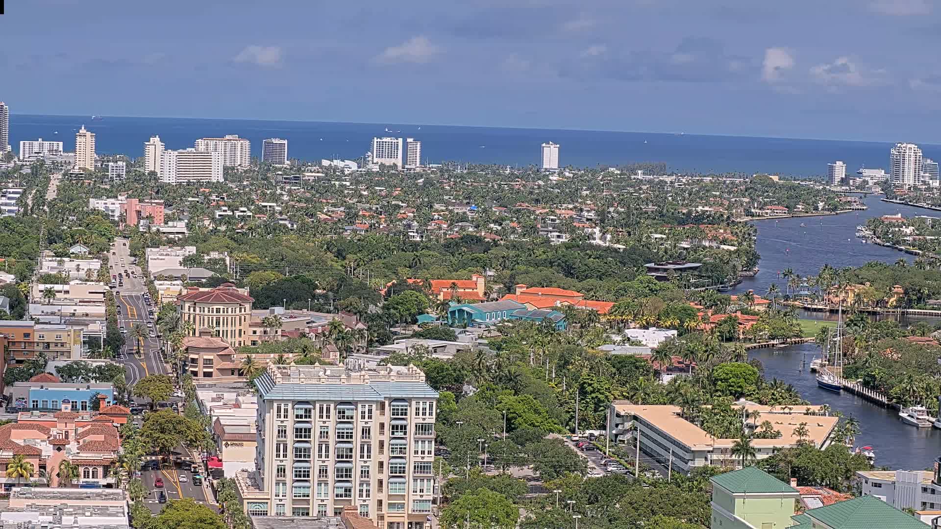 A coastal city with high-rise buildings, waterways, and lush greenery under a mostly sunny sky.