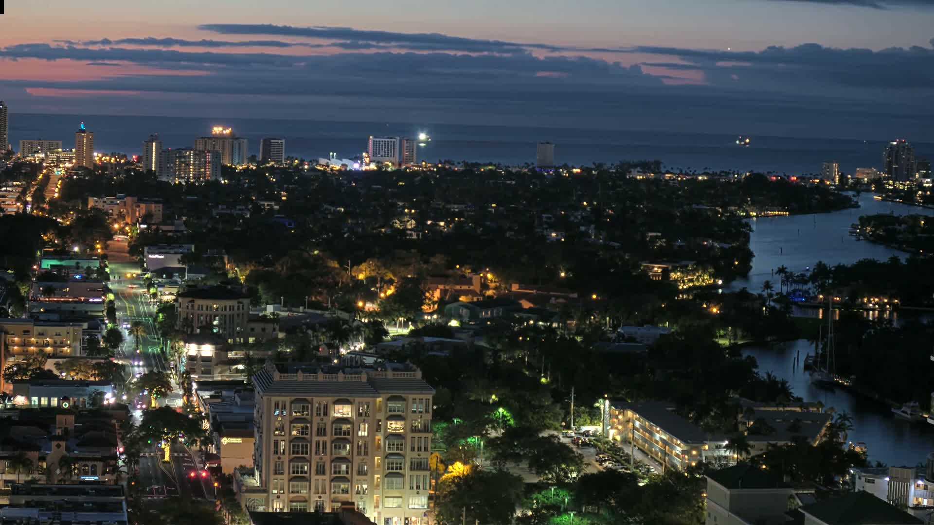 A nighttime aerial view of a city with a waterway, illuminated buildings, and a dark ocean in the background under a partly cloudy sky.