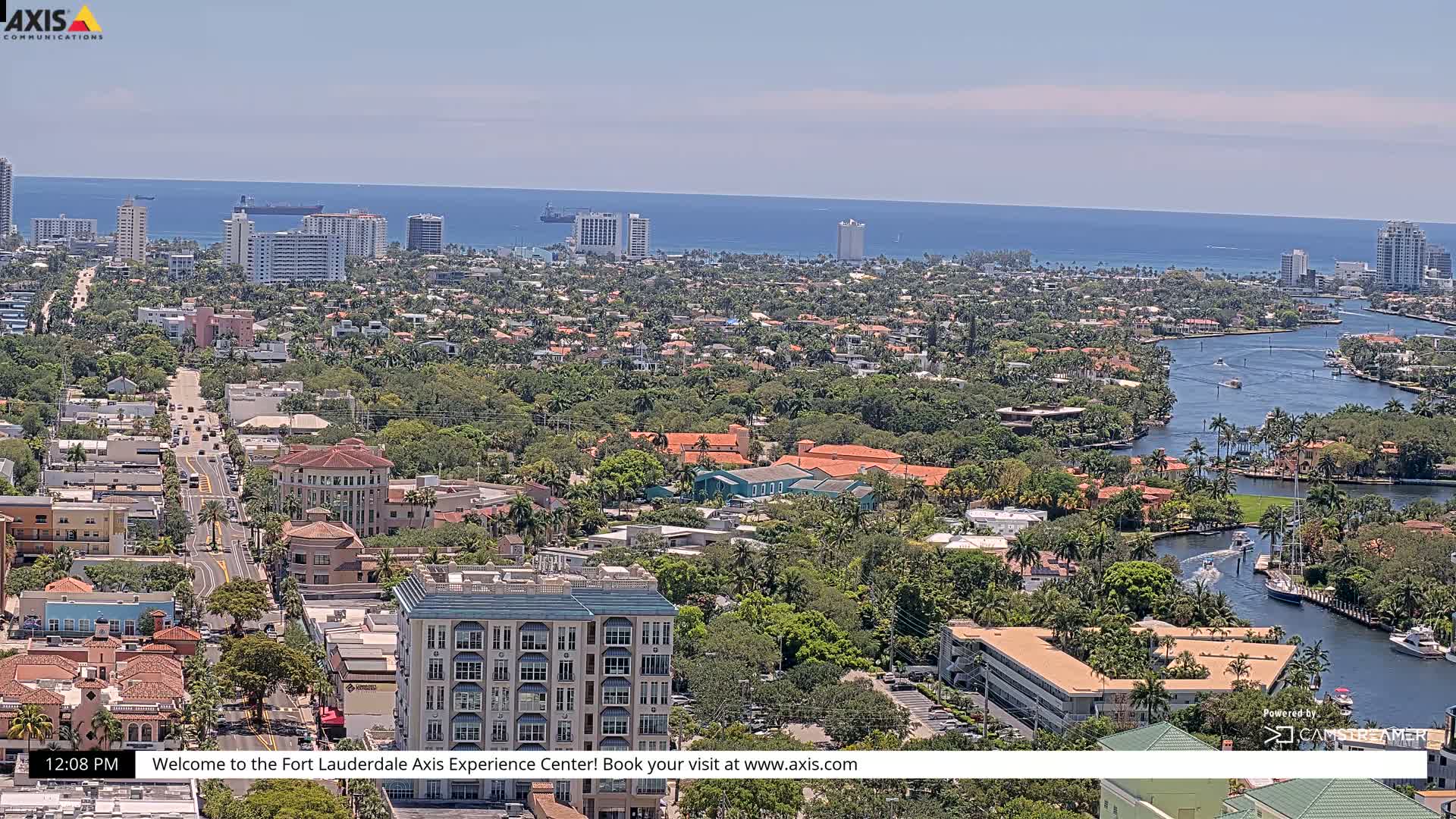 A high-angle view of a coastal city on a partly sunny day, showing a waterway winding through lush, green neighborhoods towards the ocean.