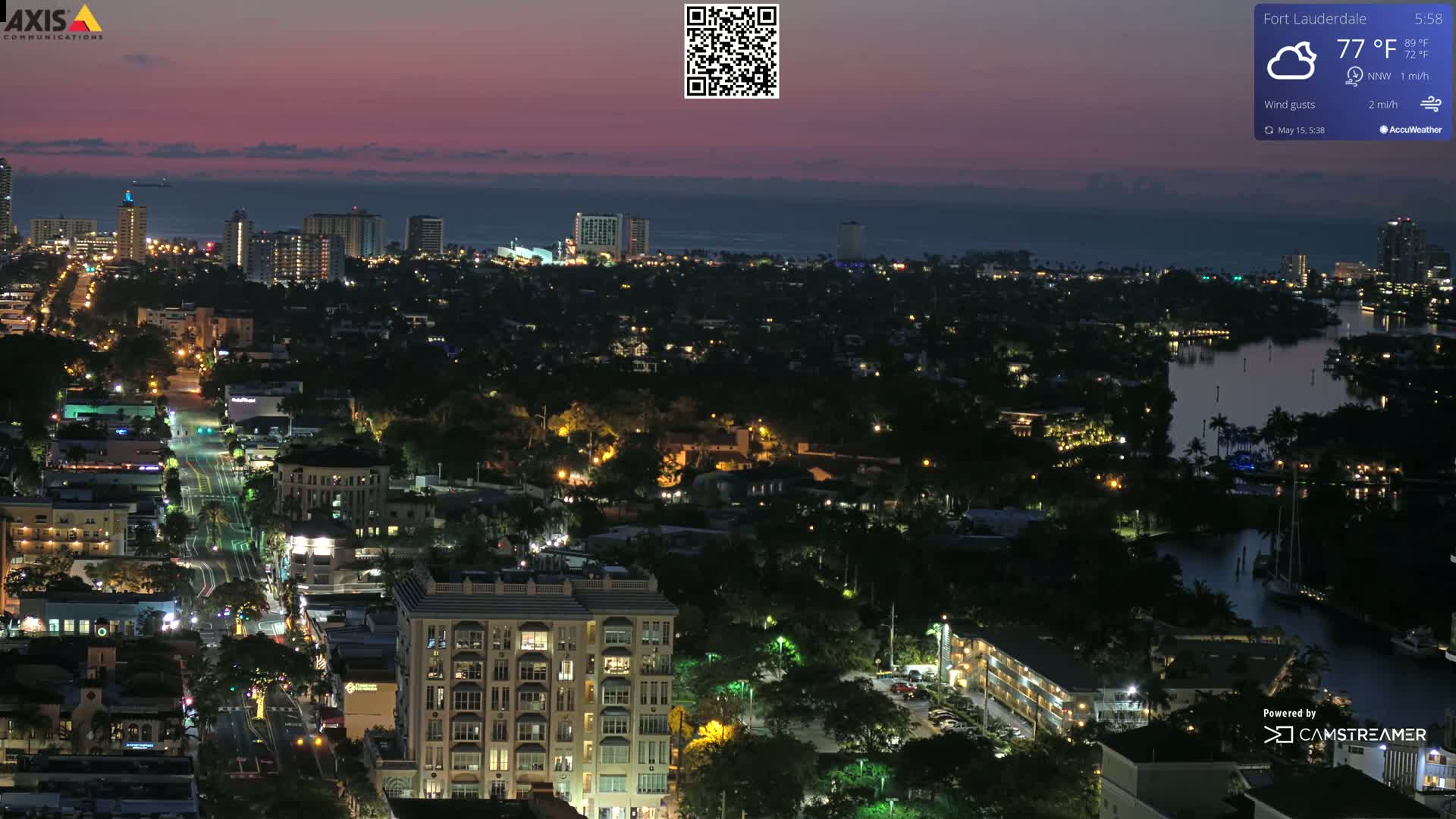 A nighttime aerial view of a city illuminated by streetlights, with a calm body of water visible in the distance under a twilight sky.