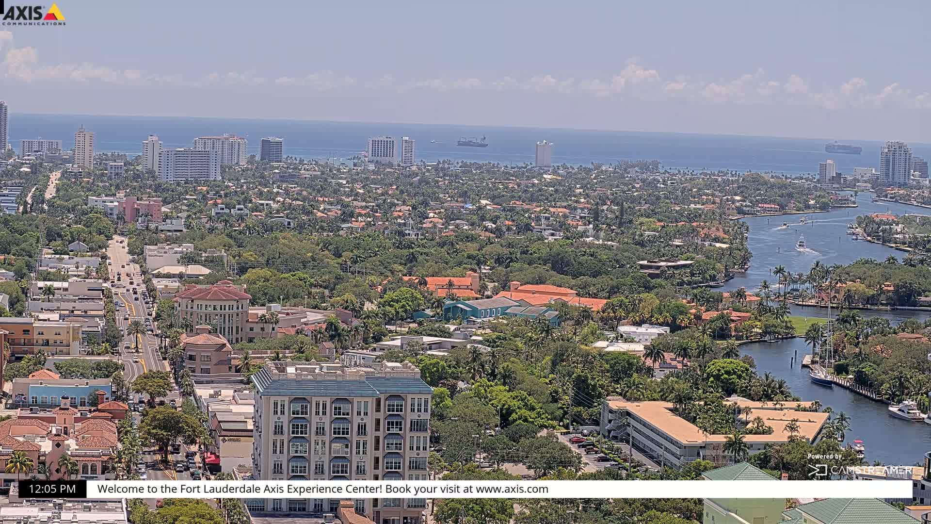 A high-angle, sunny view of a coastal city with a waterway winding through lush, green neighborhoods and high-rise buildings, extending to a calm ocean in the distance.
