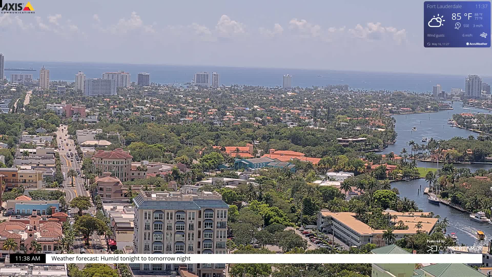 A high-angle, long shot of a coastal city on a partly sunny day with humid conditions shows a waterway winding through a residential area with many palm trees and buildings, some high-rises, extending to a calm ocean in the background.