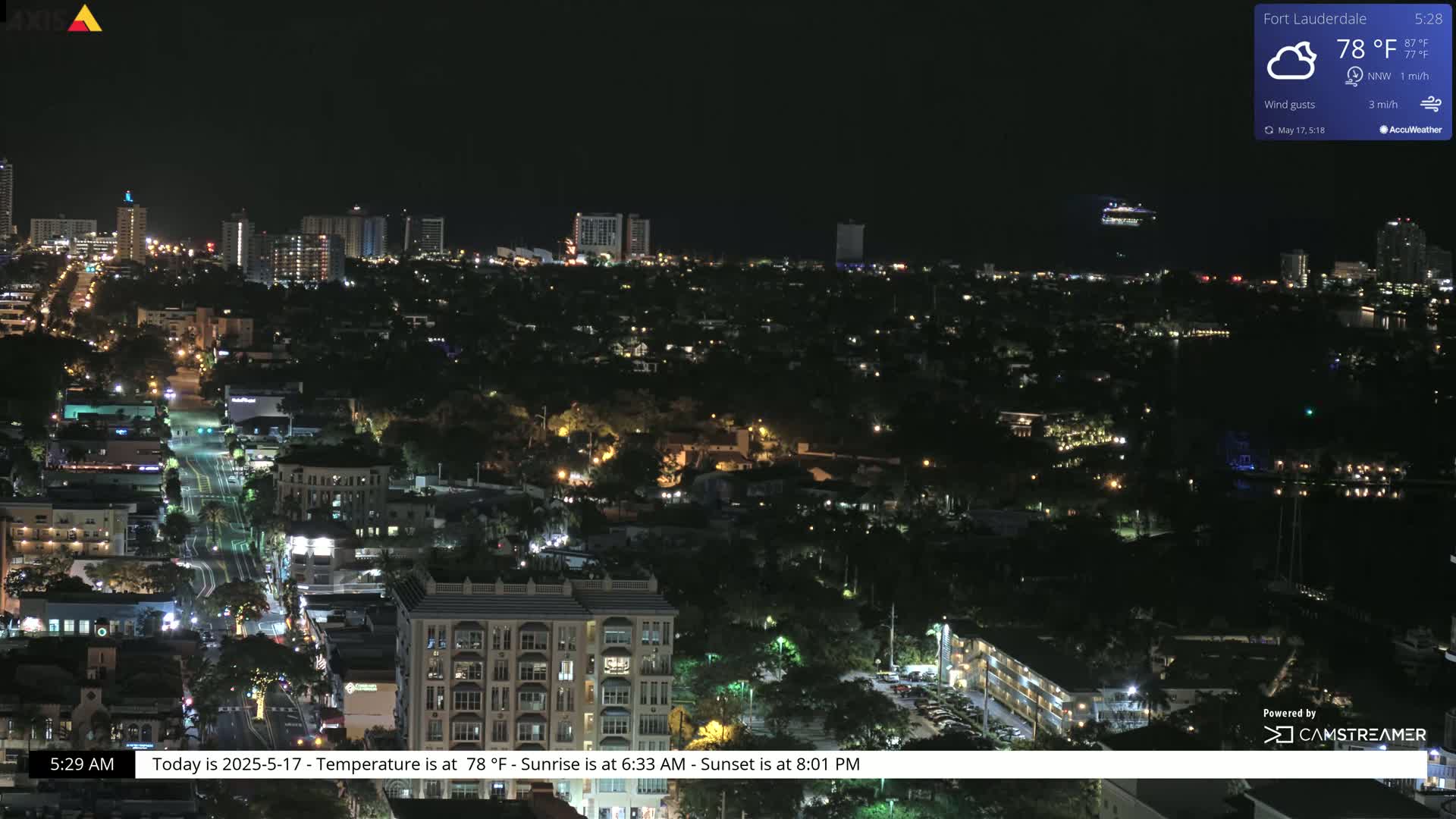 A nighttime aerial view of a city with scattered lights, a cruise ship in the distance, and partly cloudy skies.
