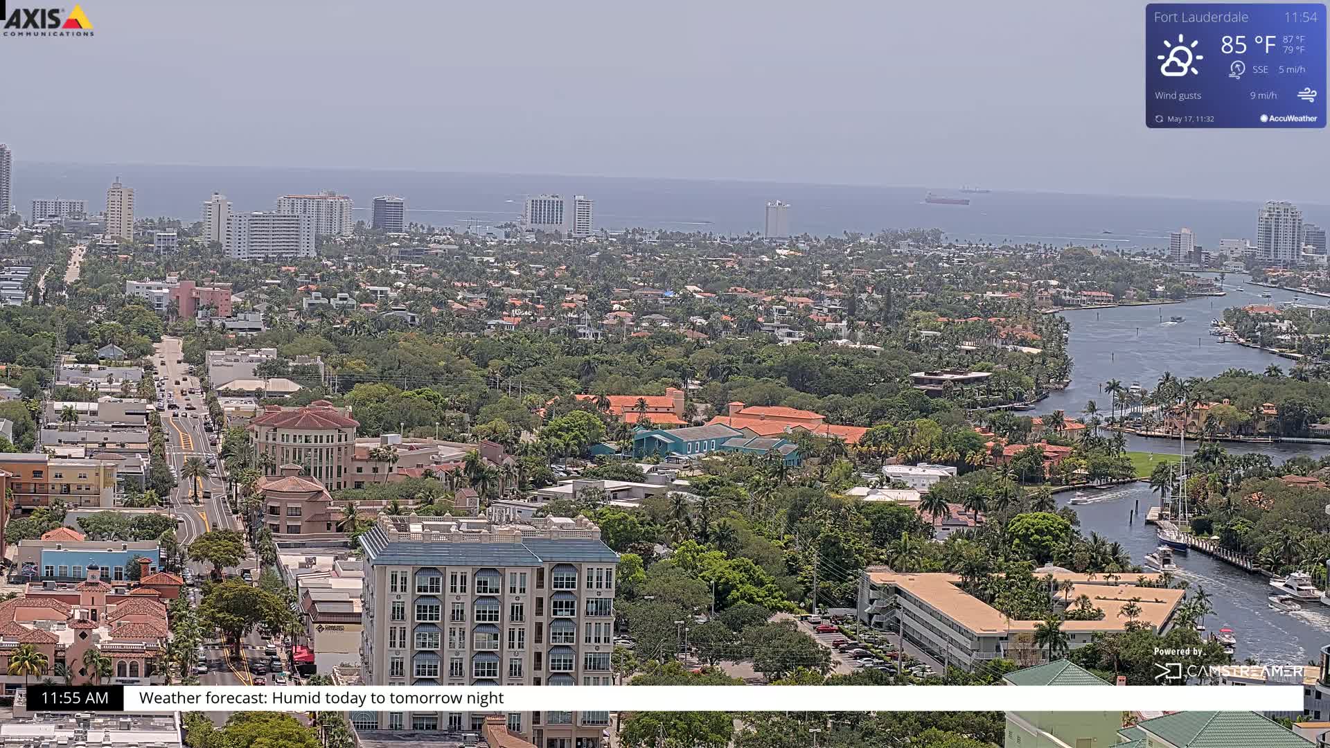 A high-angle, wide shot of a coastal city on a hazy, humid day, showing a waterway winding through a residential area towards the ocean.
