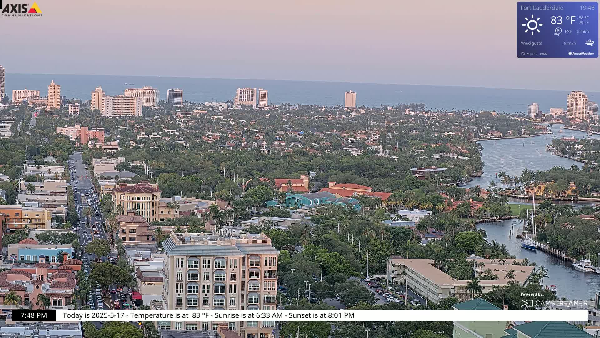 An aerial view of a coastal city at dusk shows a calm ocean, a network of waterways, and numerous buildings, under a clear sky.