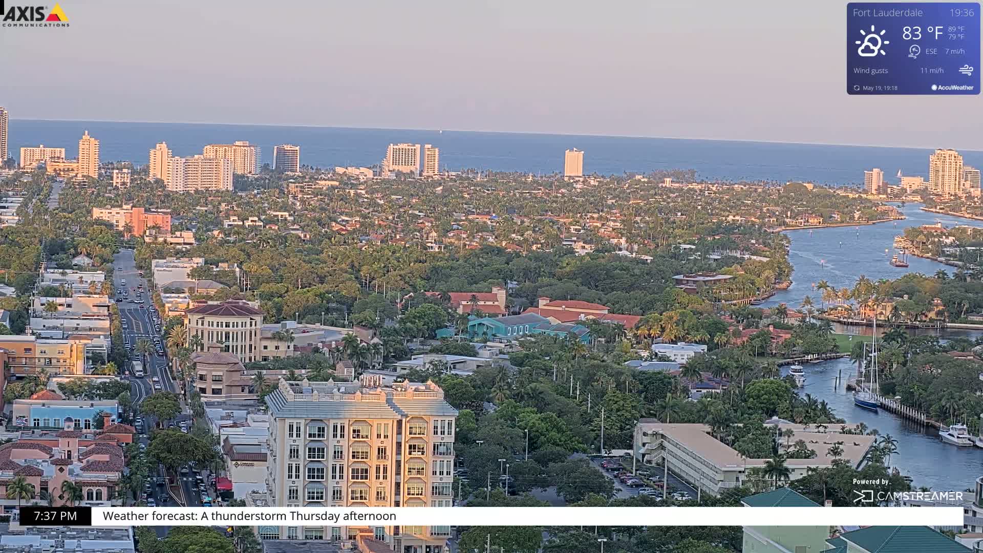 An aerial view of a coastal city at sunset, showing a waterway winding through a residential area with buildings and palm trees, under partly cloudy skies.