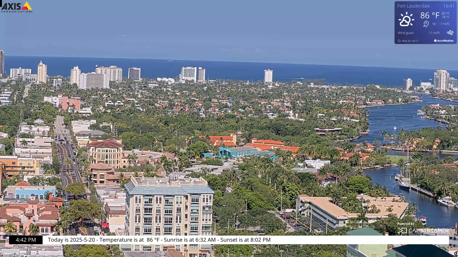 An aerial view shows a coastal city with buildings, palm trees, and waterways under sunny skies.